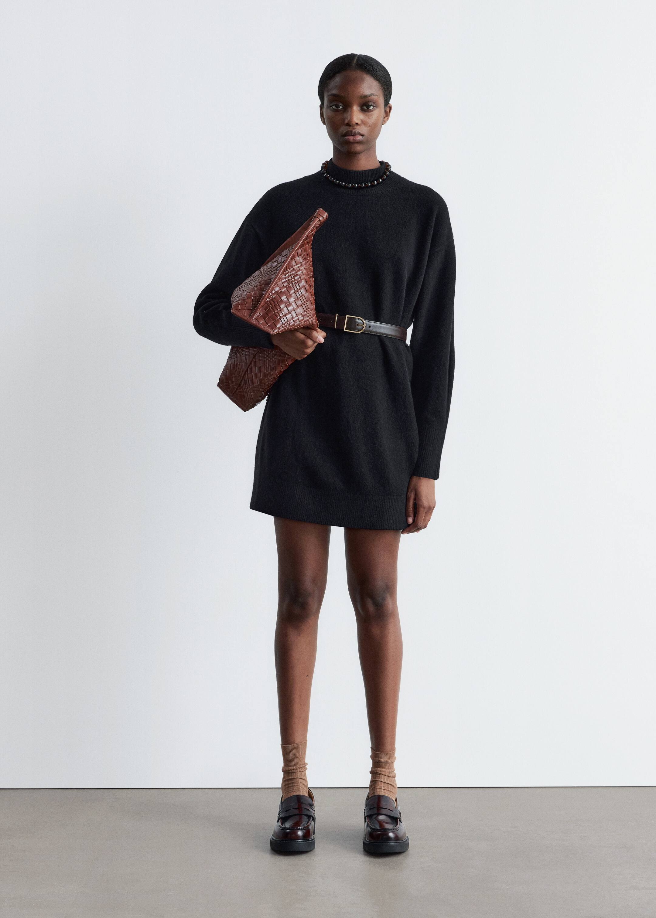 Größeres Bild anzeigen: Das Modell trägt ein schwarzes, entspanntes Mini-Strickkleid mit Stehkragen und langen Ärmeln, das durch einen tonalen Schnallengürtel in der Taille betont wird.
