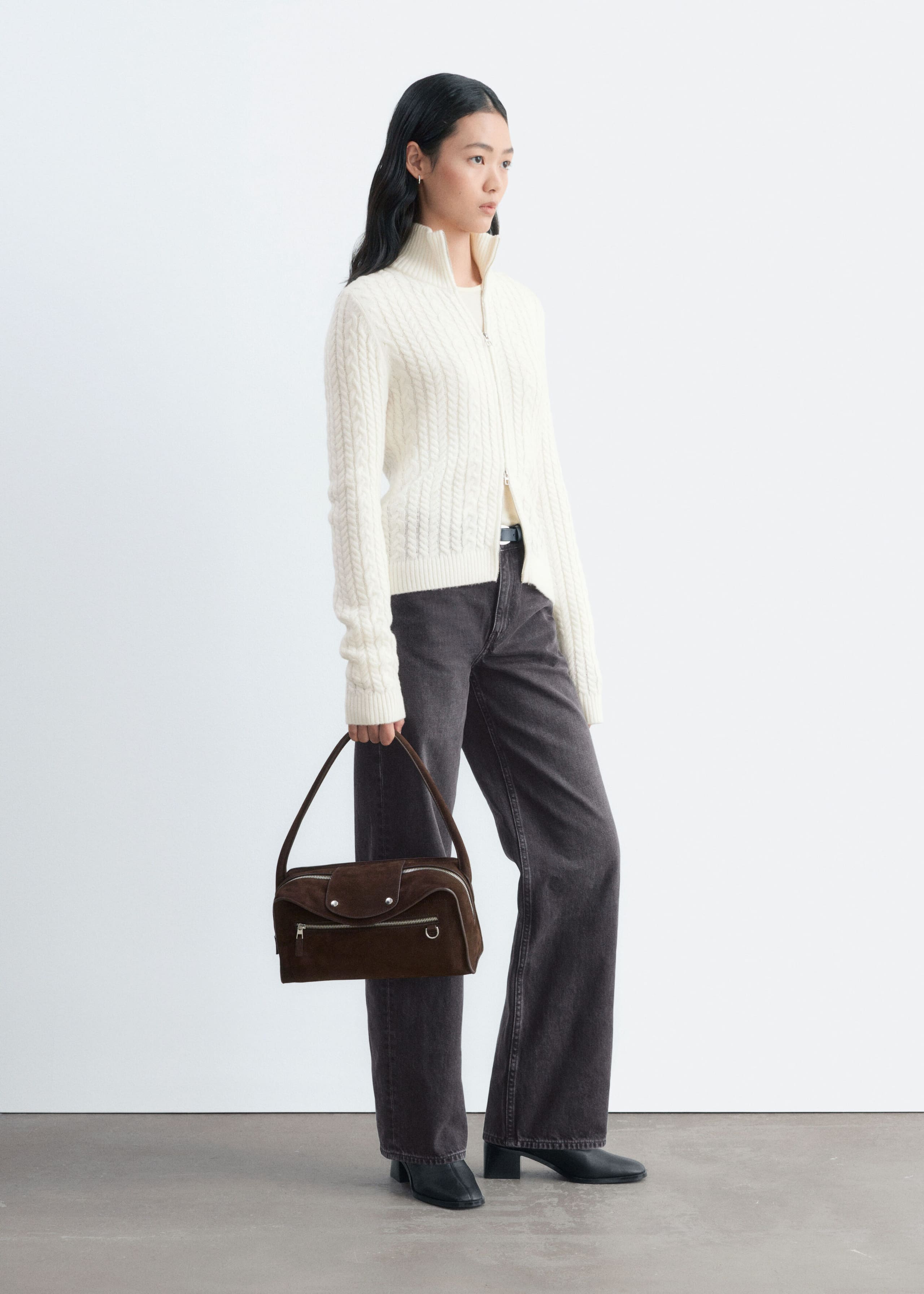 A cream-coloured, slim-fit cable-knit cardigan is being worn by the model, featuring a front zip and a high ribbed collar. The cardigan is styled with dark grey wide-leg jeans, and the model is holding a brown suede handbag.
