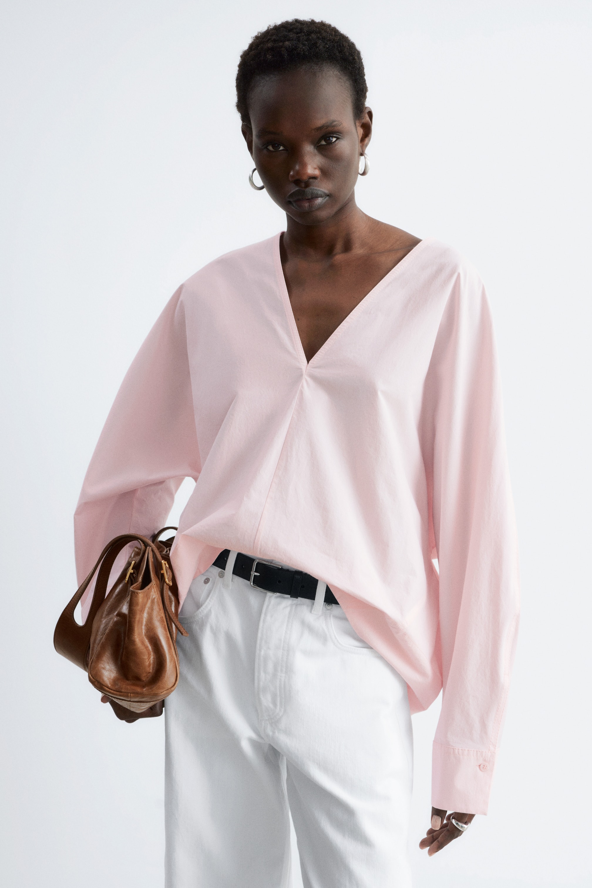 Grösseres Bild ansehen: Eine hellrosa, oversized Langarmbluse mit tiefem V-Ausschnitt und überschnittenen Schultern, sowie geknöpften Manschetten. Die Bluse ist teilweise in weiße Hosen gesteckt und zeigt einen schwarzen Gürtel.