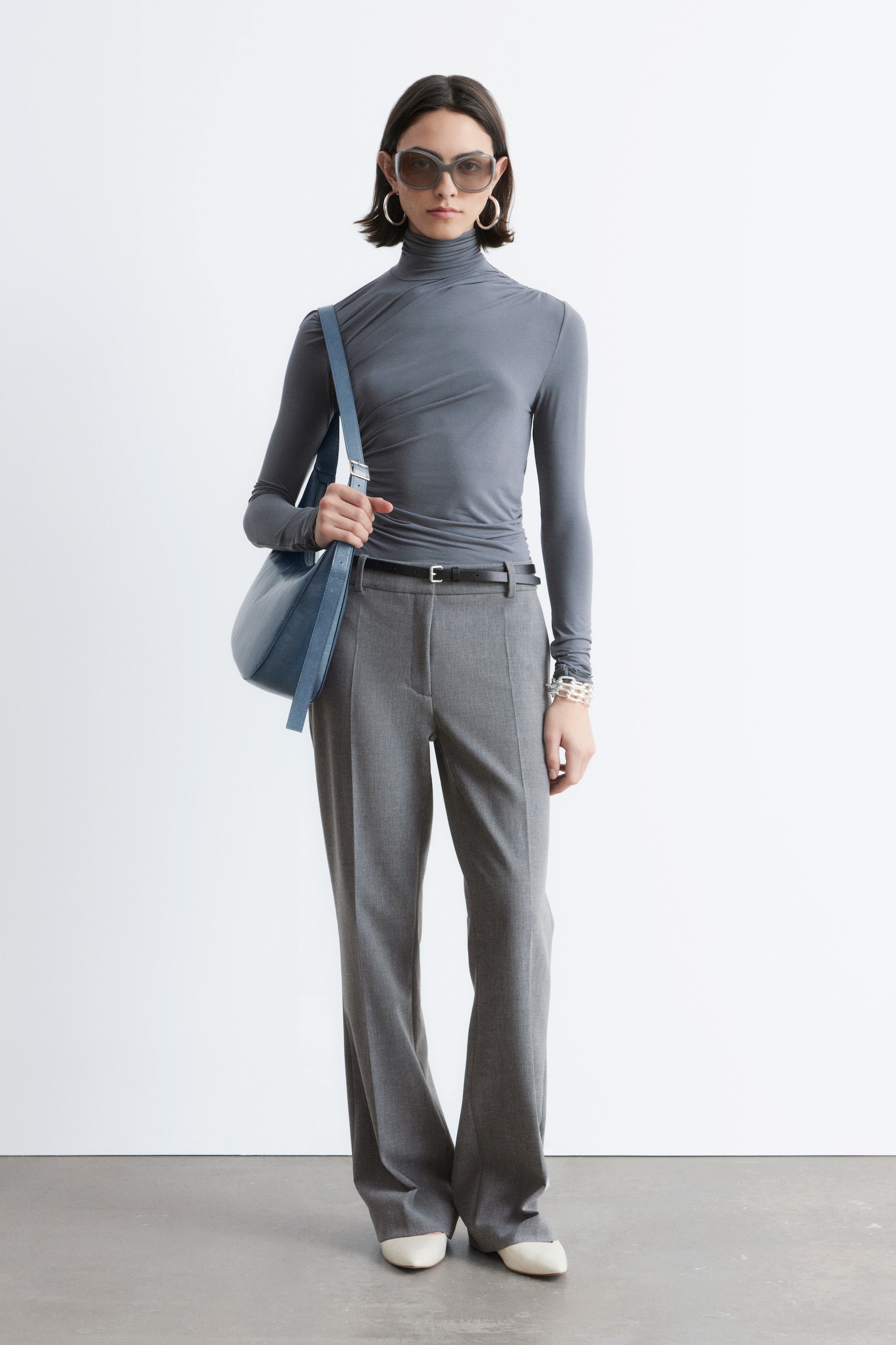 Näytä suurempi kuva: Tumma harmaa, pitkähihainen poolopaita, jossa on hienovaraisia laskoksia kaulan ja vartalon ympärillä, yhdistetään vaaleanharmaisiin, korkeavyötäröisiin leveälahkeisiin housuihin, joita korostaa ohut musta vyö. Asukokonaisuuden täydentävät vaalean kermanväriset, teräväkärkiset ballerinat ja vaaleansininen, rennosti olalla kannettava olkalaukku.