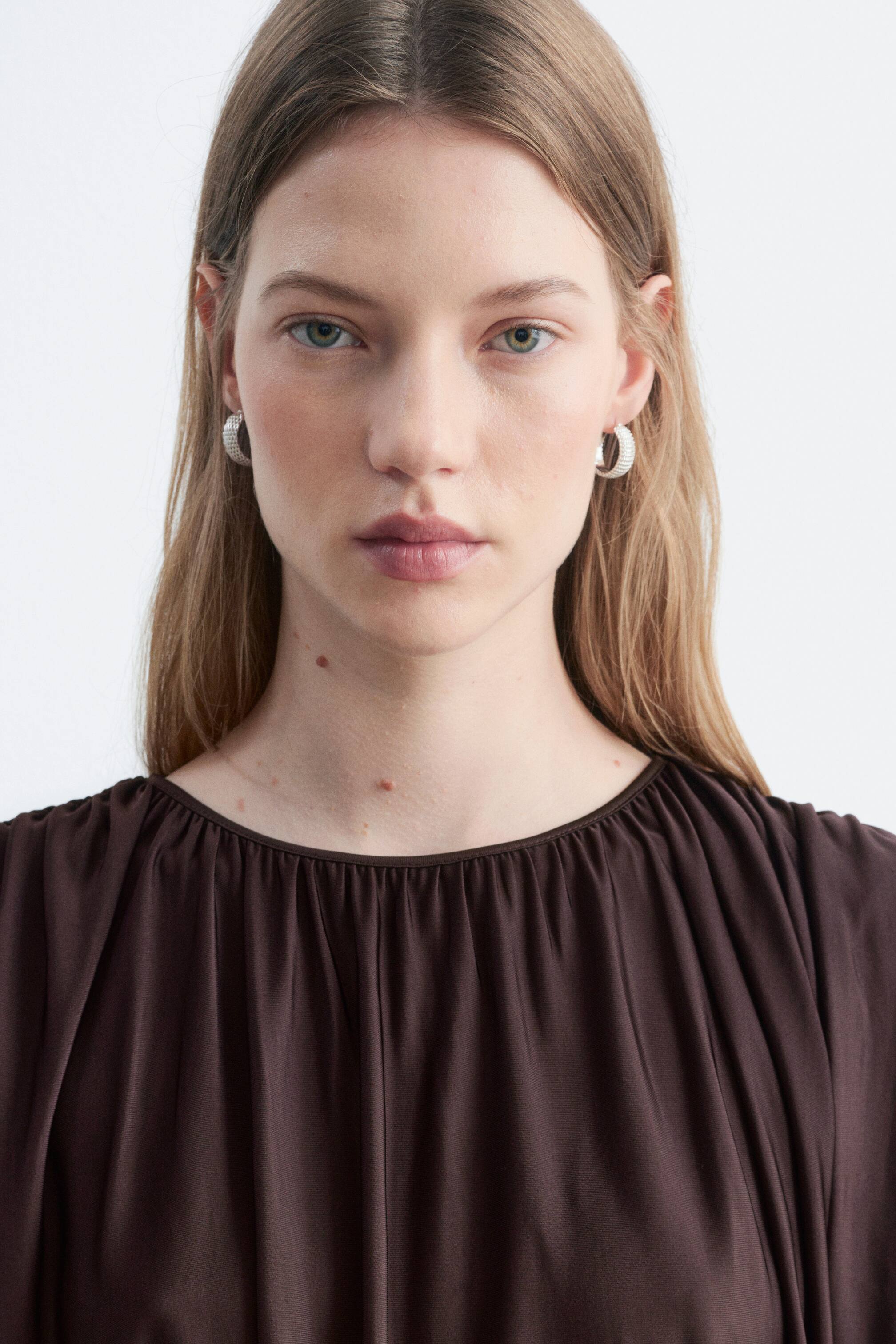 Grotere afbeelding bekijken: Een donkerbruine mouwloze jersey top, met zachte, volumineuze drapering rond de geplooide ronde hals, wordt gedragen door het model.