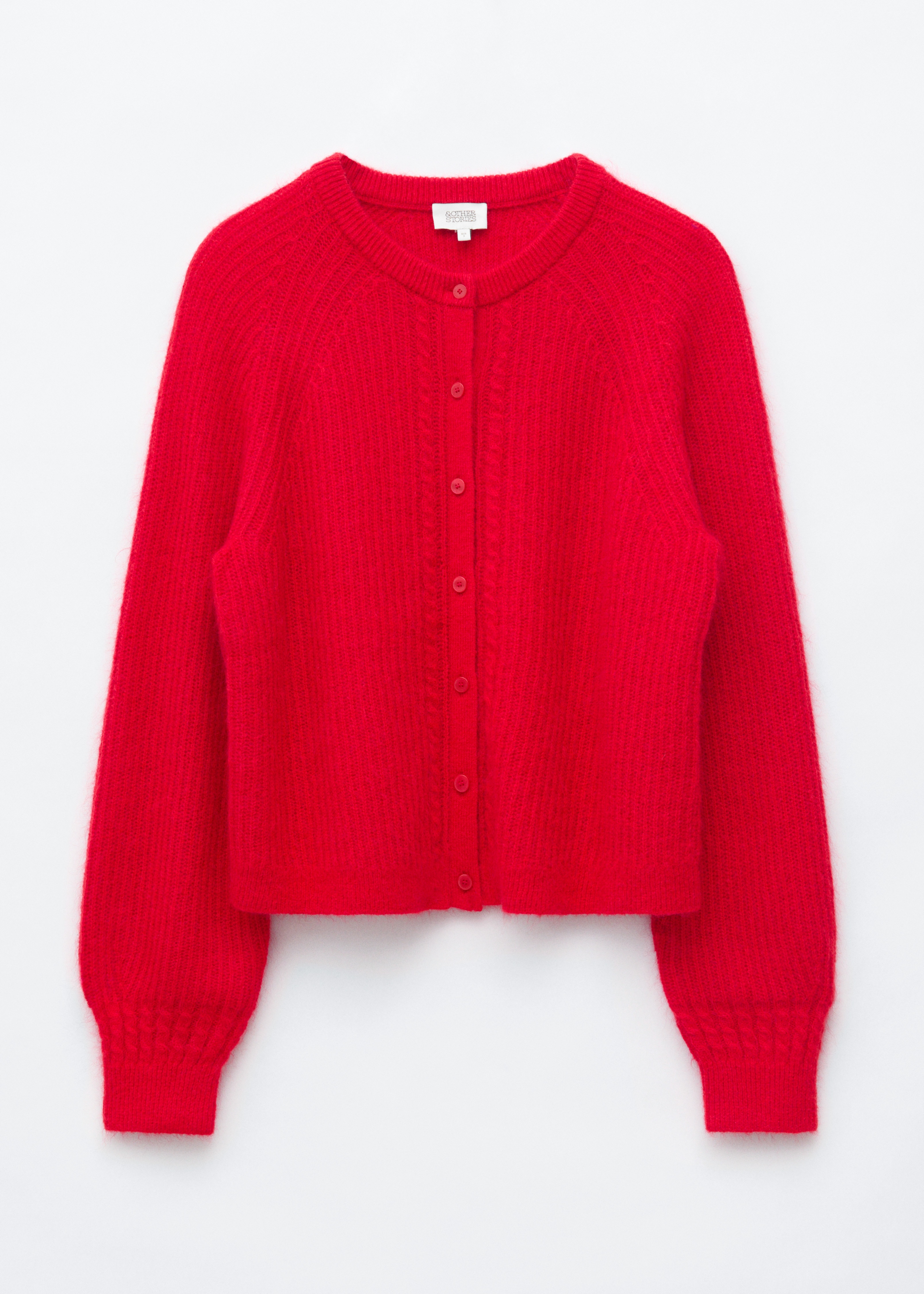 La modella indossa un cardigan rosso testurizzato con lavorazione a maglia a coste e dettagli a trecce lungo lâabbottonatura. Il cardigan Ã¨ indossato con il collo parzialmente sbottonato, mettendo in risalto una sottile collana a catena dorata.