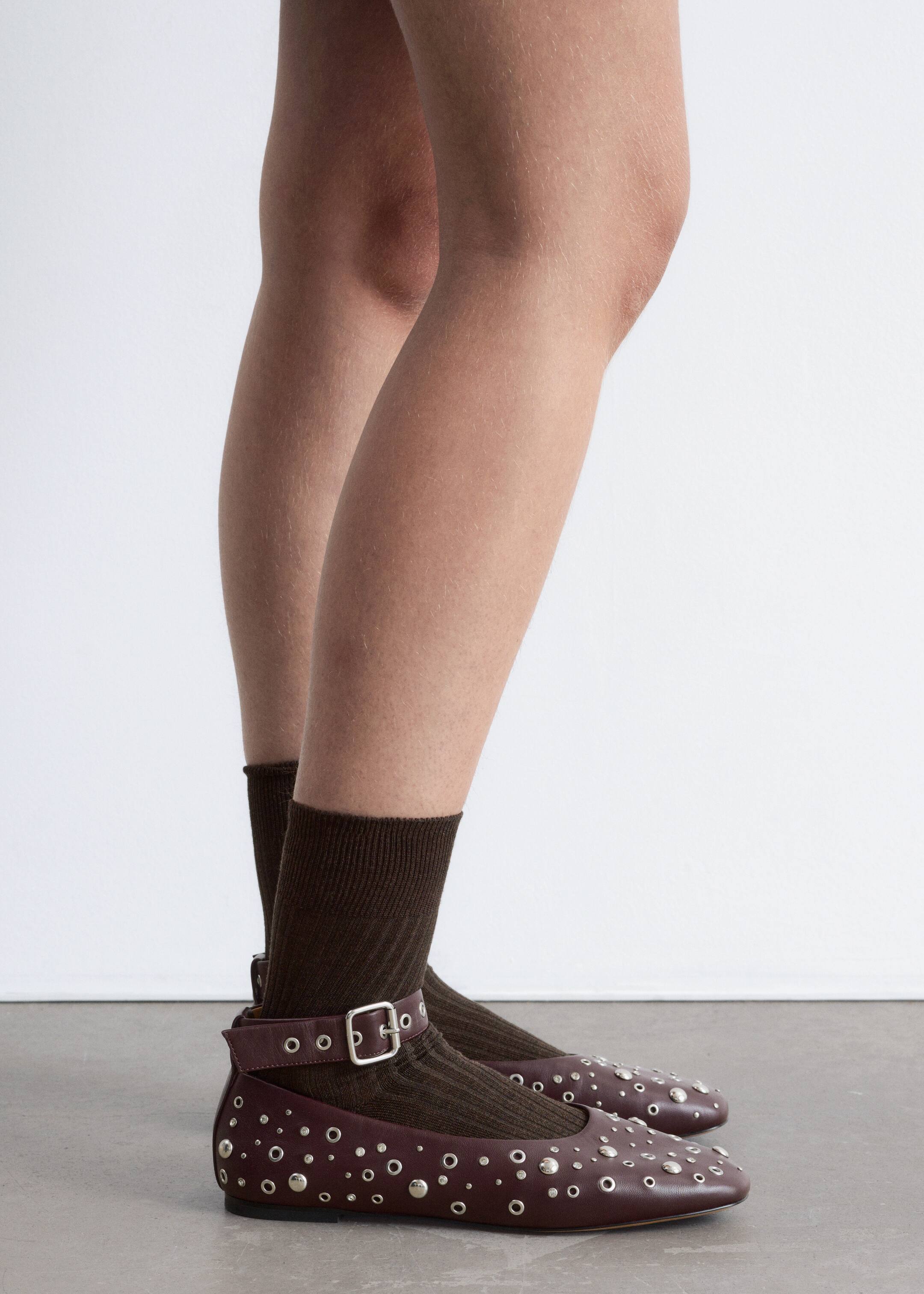 View larger image: Dark burgundy leather flats are being worn, seen from a side profile. They are featuring an ankle strap with a silver buckle, adorned with silver studs and eyelets across the vamp and sides, and are layered over dark brown ribbed socks.