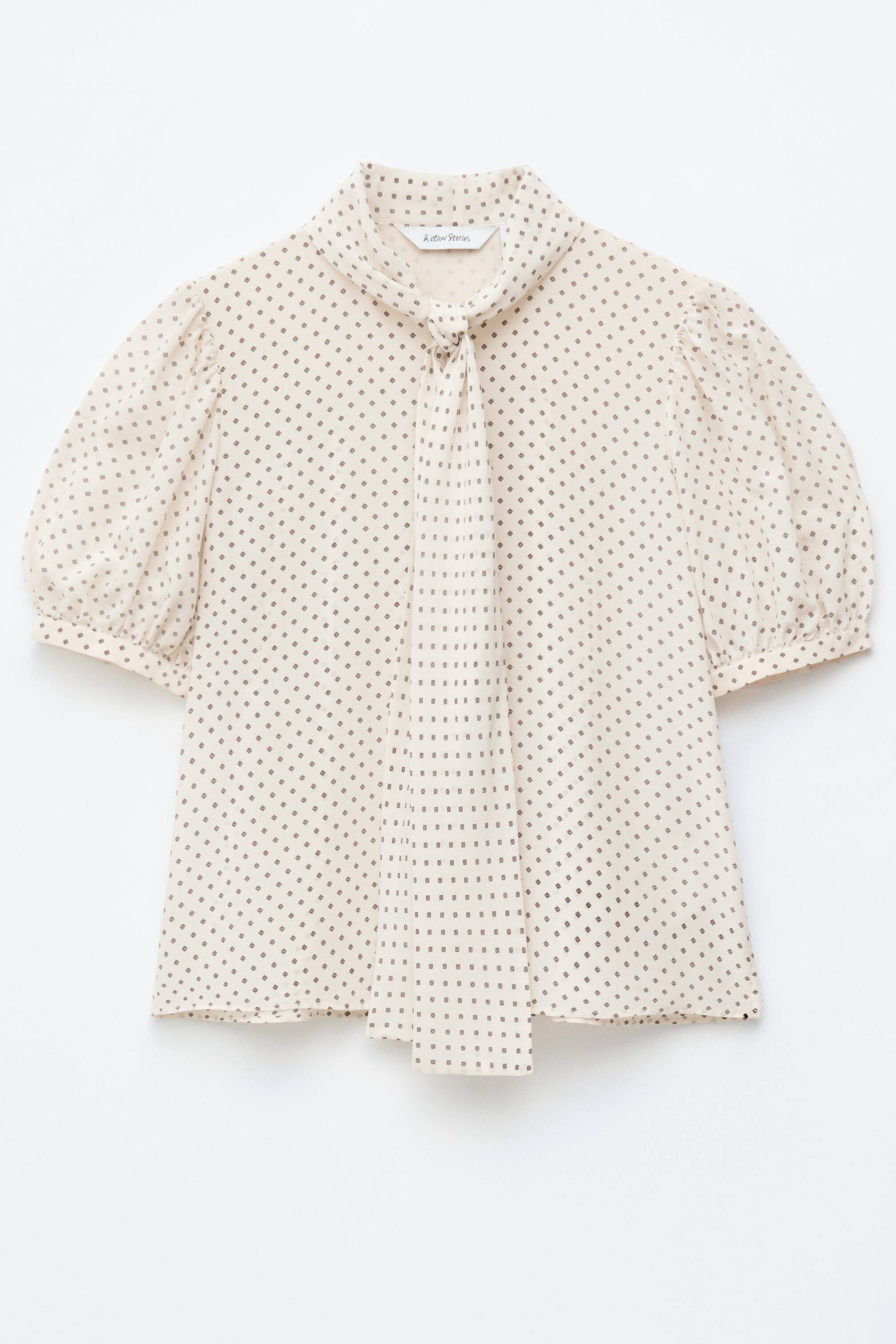 Agrandir l'image: Le DEVANT d'un chemisier crème présente un motif micro intégral de petits carrés foncés. Il est conçu avec de délicates manches bouffantes froncées aux épaules et aux poignets, ainsi qu'un col à nouer qui se ferme à l'avant.