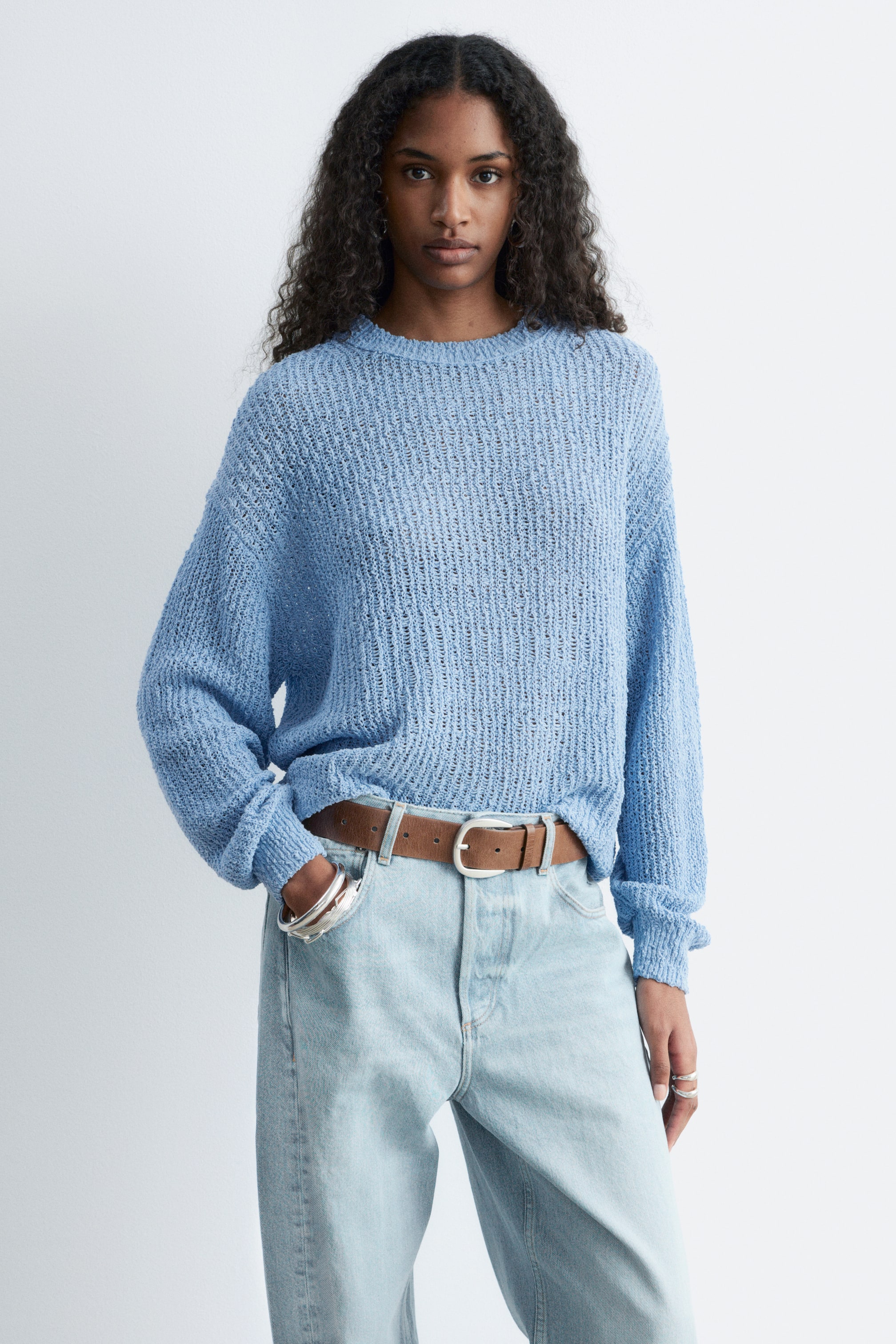 Se større billede: En lyseblå åbenstrikket sweater præsenteres forfra, med rund hals, lange ærmer og en afslappet pasform, hvor kanten delvist er stukket ned i jeans.