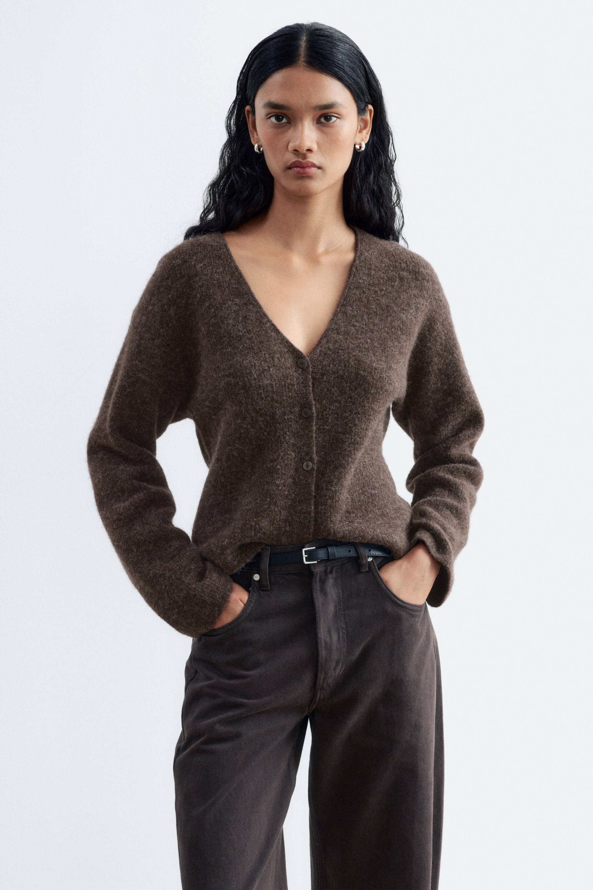Grösseres Bild ansehen: Ein brauner Strick-Cardigan mit Struktur, V-Ausschnitt und Knopfleiste wird vom Model getragen. Der Cardigan ist mit dunklen Hosen und einem schmalen Gürtel kombiniert, wobei das Model die Hände in den Taschen hält.