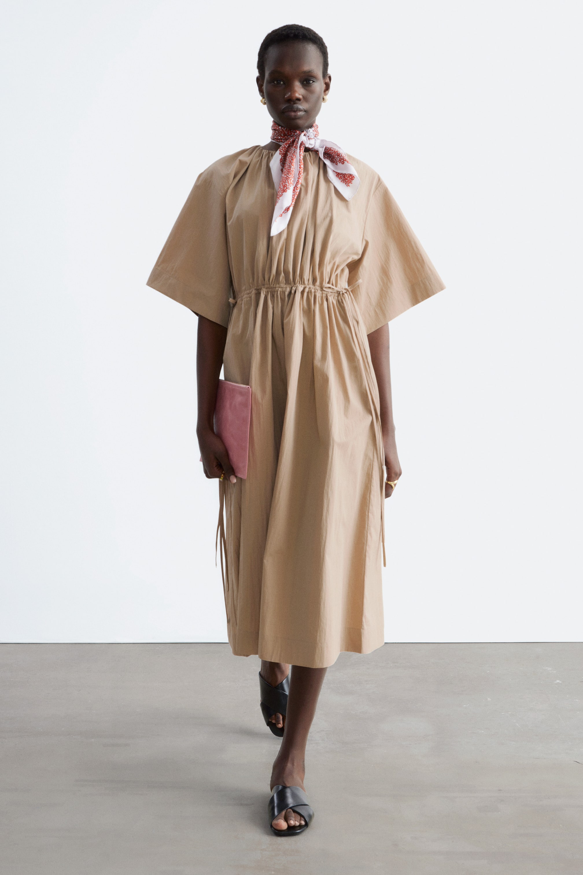 Ver imagem maior: Um vestido midi bege exibe um decote redondo alto franzido e mangas curtas largas. É ajustado na cintura por um cordão, criando franzidos, e flui para uma saia completa que chega abaixo do joelho.