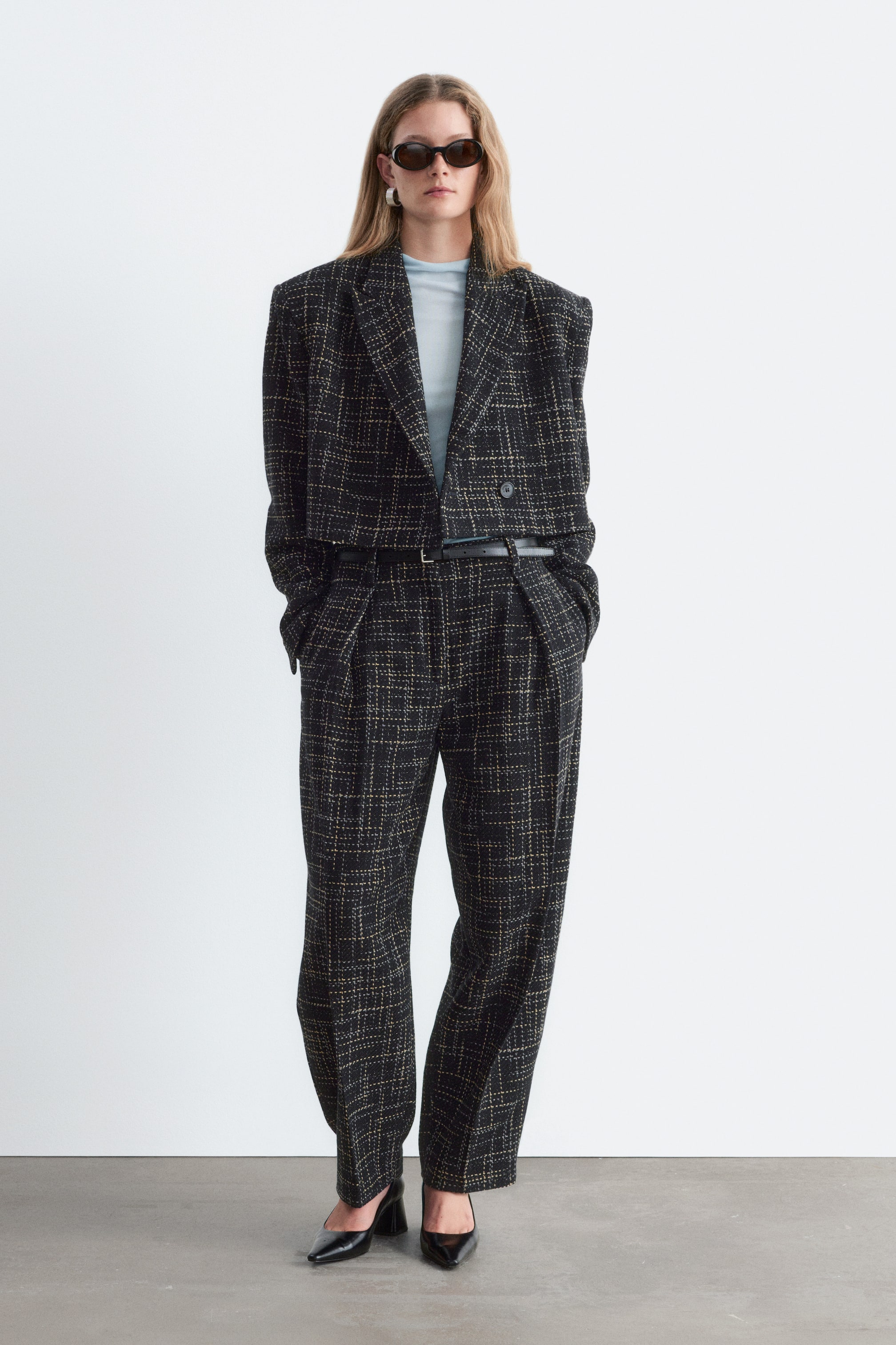 Größeres Bild ansehen: Ein verkürzter Blazer mit einem strukturierten Karomuster in Schwarz und Gelb ist von vorne zu sehen, mit fallenden Revers und formgebenden Schulterpolstern. Der Blazer in Boxy-Passform wird über einem hellblauen Oberteil getragen und mit einer passenden Hose kombiniert, um die verkürzte Länge hervorzuheben.