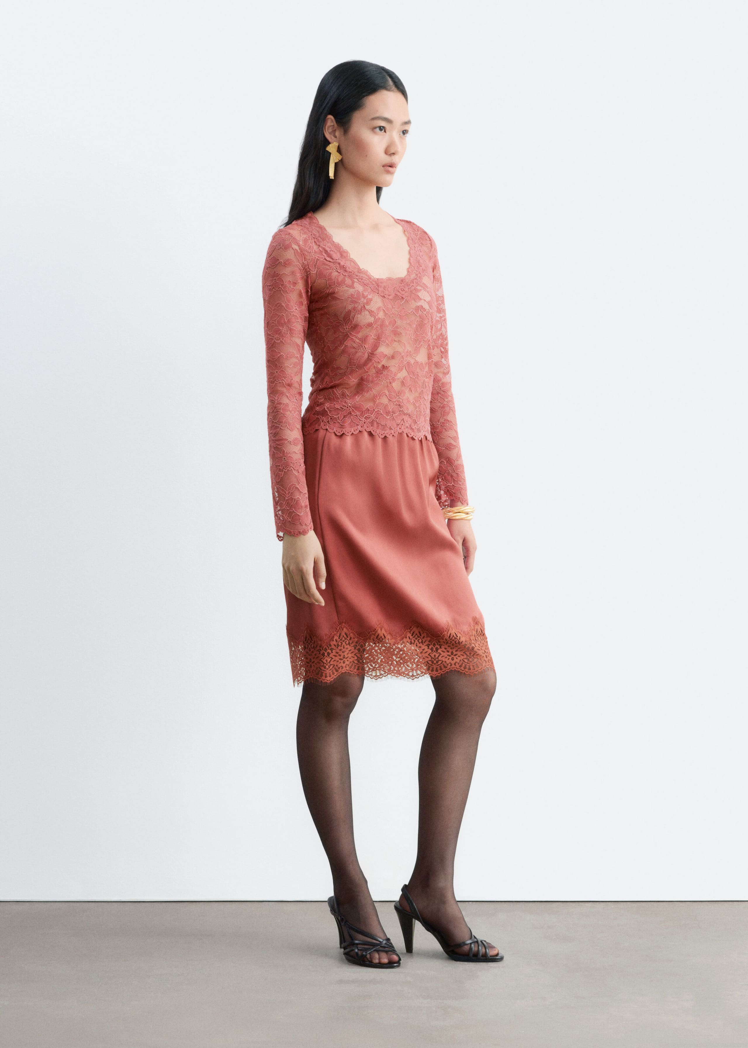 The model is wearing a long-sleeved dusty rose lace top and a matching satin knee-length skirt. The slim-fit top features a scoop neckline and scalloped edges, while the skirt is finished with a delicate lace-trimmed hem. The outfit is styled with sheer black tights and strappy black heels.