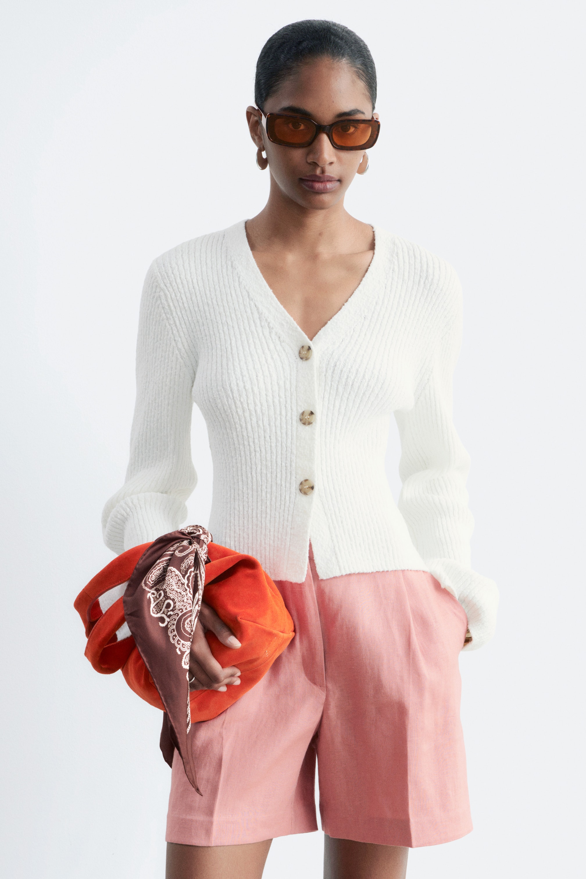 Zobacz większy obraz: Modelka ma na sobie biały prążkowany kardigan z dekoltem w serek i jasnymi guzikami. Lekko skrócony kardigan charakteryzuje się dopasowanym krojem i jest zestawiony z jasnoróżowymi szortami.