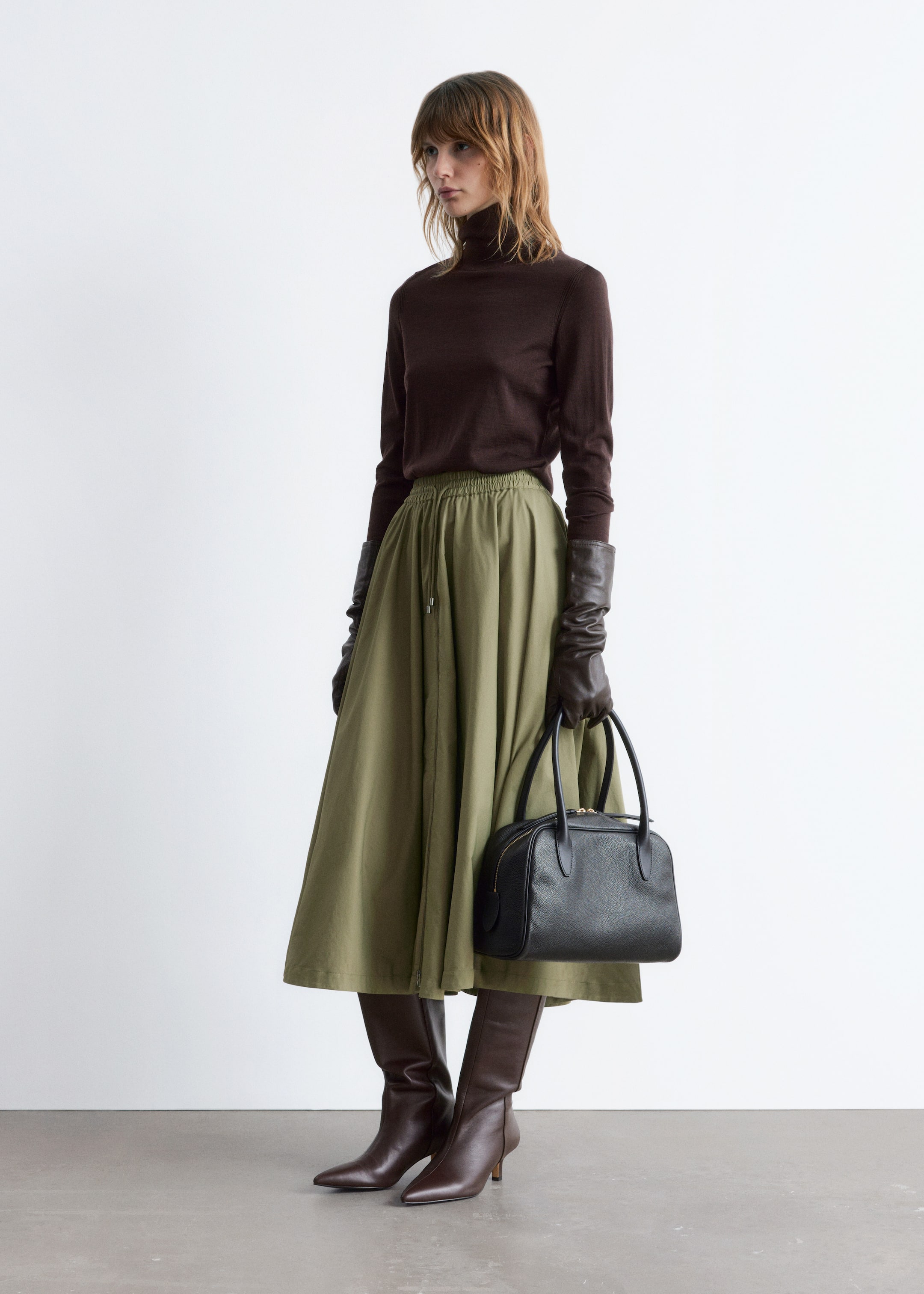 Visa större bild: En modell visar upp en outfit från en trekvarts frontvy, iklädd en mörkbrun långärmad polotröja och en olivgrön hel midikjol med en rynkad elastisk midja med ett synligt dragsko. Hon har accessoarer som består av mörkbruna långa läderhandskar, mörkbruna knähöga spetsiga stövlar och en svart strukturerad handväska med dubbla topphandtag och en synlig dragkedjestängning.
