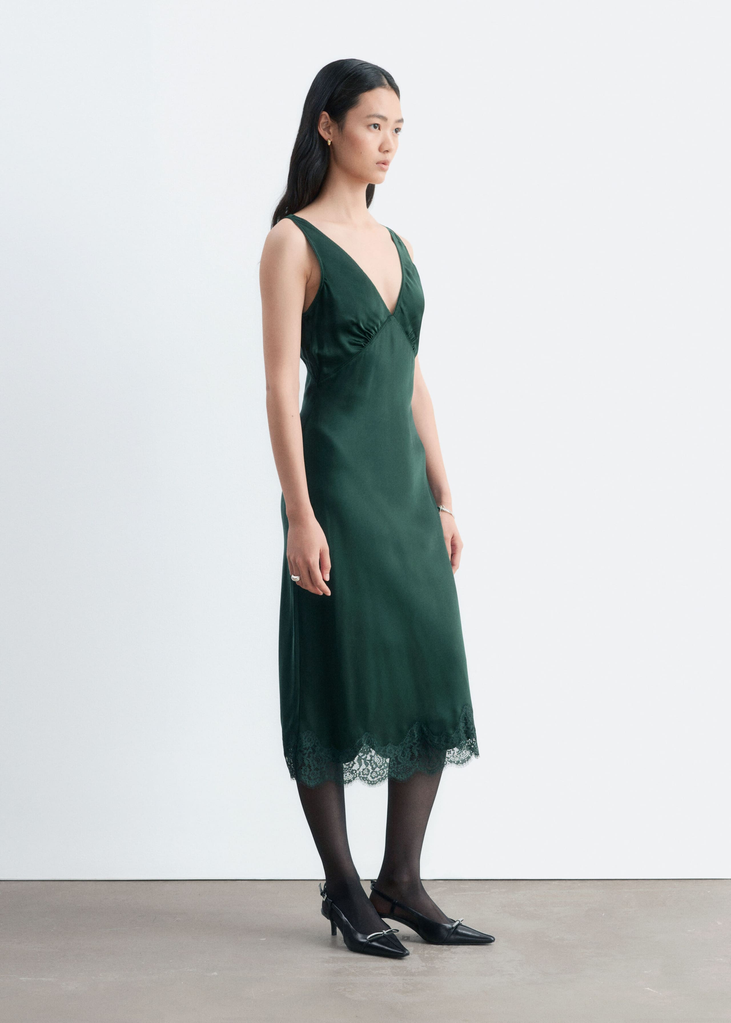A dark green slip dress featuring a V-neckline, gathered bust, and scalloped lace hem is worn by the model. The dress falls to a midi length and is paired with sheer black tights and black pointed-toe heels.