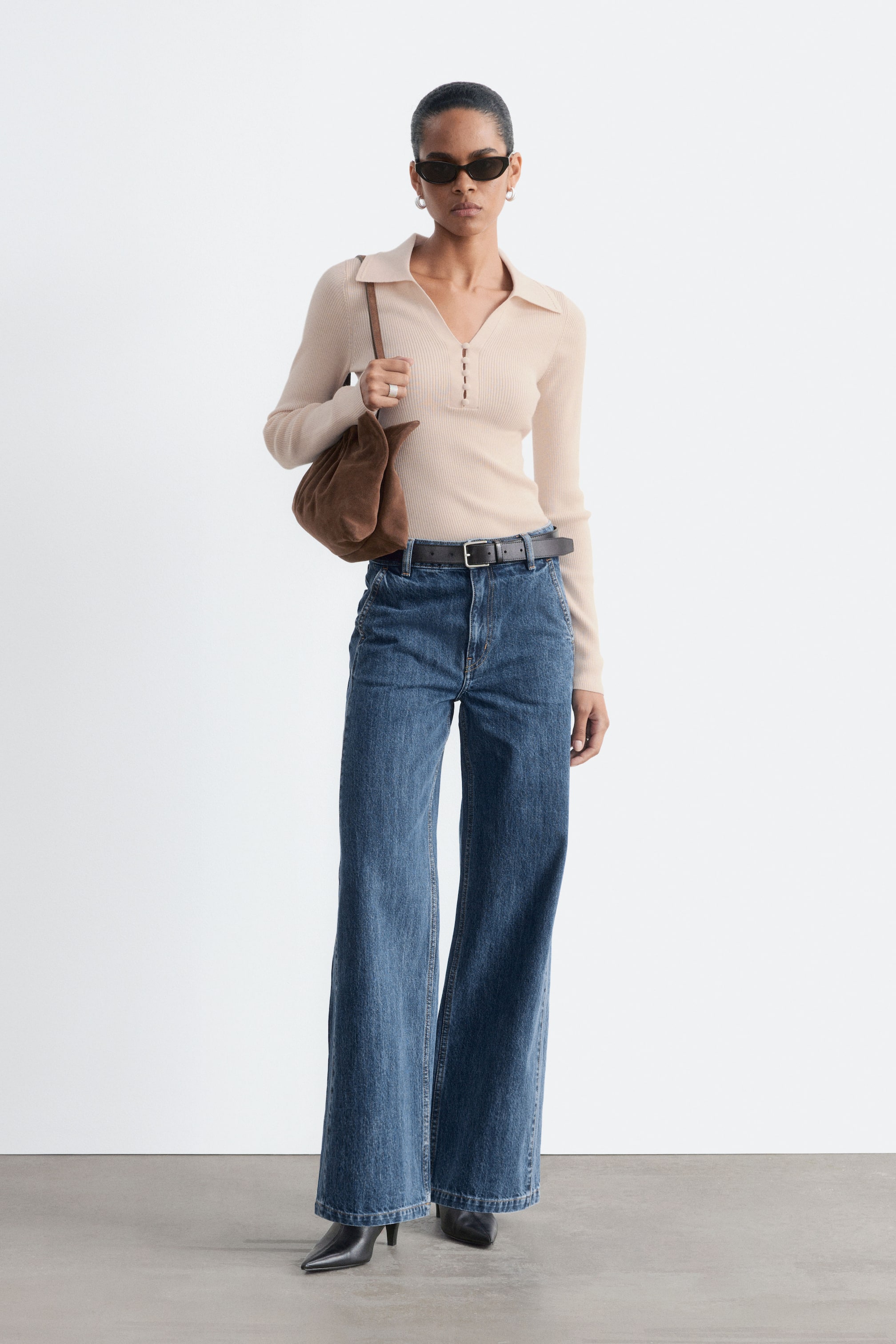 Ver imagen más grande: La modelo lleva unos vaqueros denim azules, de tiro alto y pernera ancha, combinados con un cinturón de cuero negro ceñido a la cintura. Los jeans presentan una silueta relajada con bolsillos frontales inclinados y se combinan con un top beige de manga larga y canalé, metido por dentro, y unos botines negros de punta.