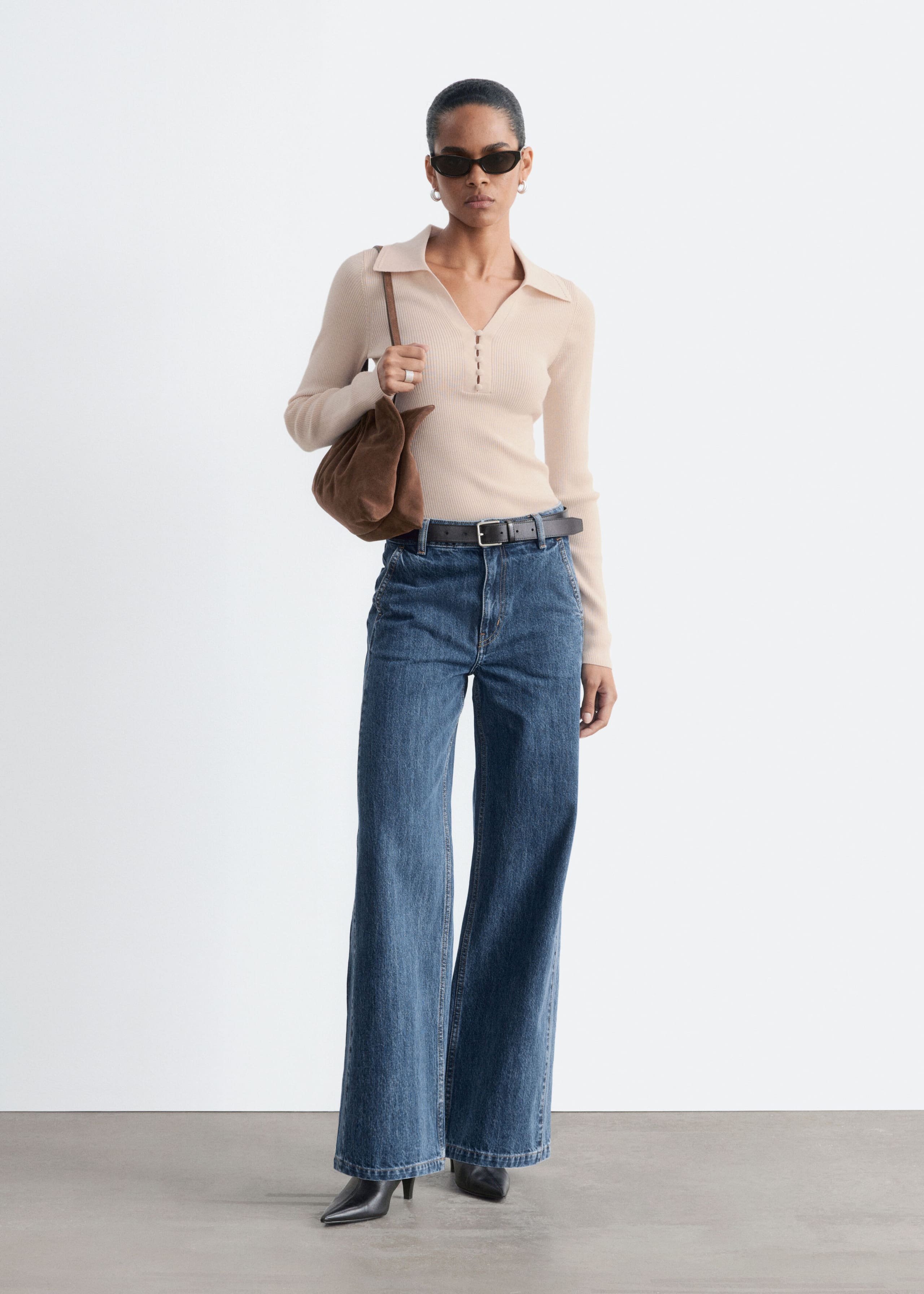 The model is wearing blue, high-waisted wide-leg denim jeans, styled with a black leather belt cinched at the waist. The jeans feature a relaxed silhouette with slanted front pockets and are paired with a tucked-in beige ribbed long-sleeve top and black pointed-toe boots.