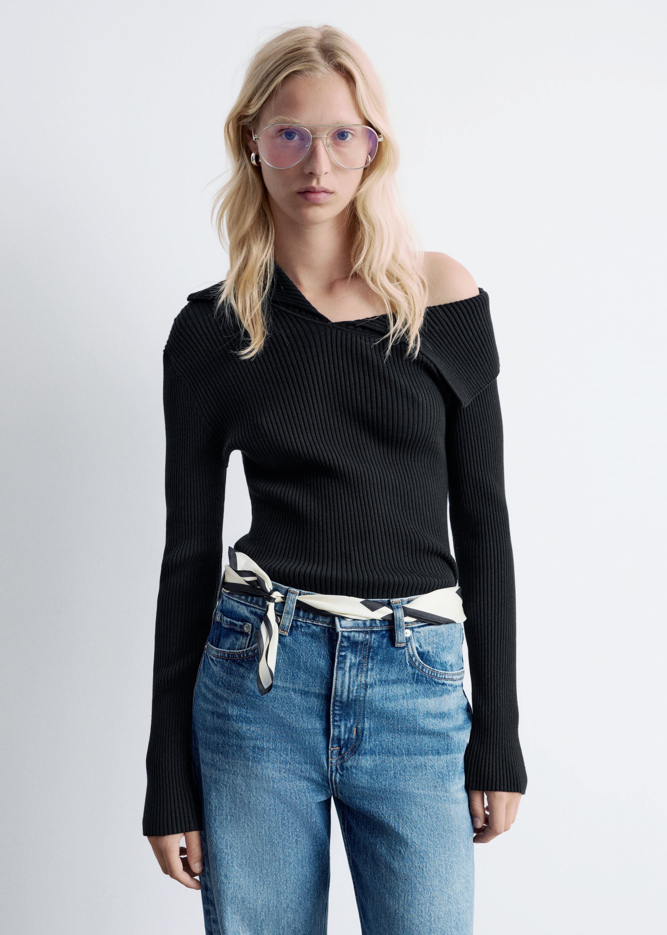 Agrandir l'image: Un haut noir en tricot côtelé à manches longues est porté par un mannequin. Le haut présente un décolleté asymétrique avec un col marin, qui est replié pour créer un style épaules dénudées d'un côté. Il est stylé avec un jean bleu et un foulard à motifs noué à la taille.