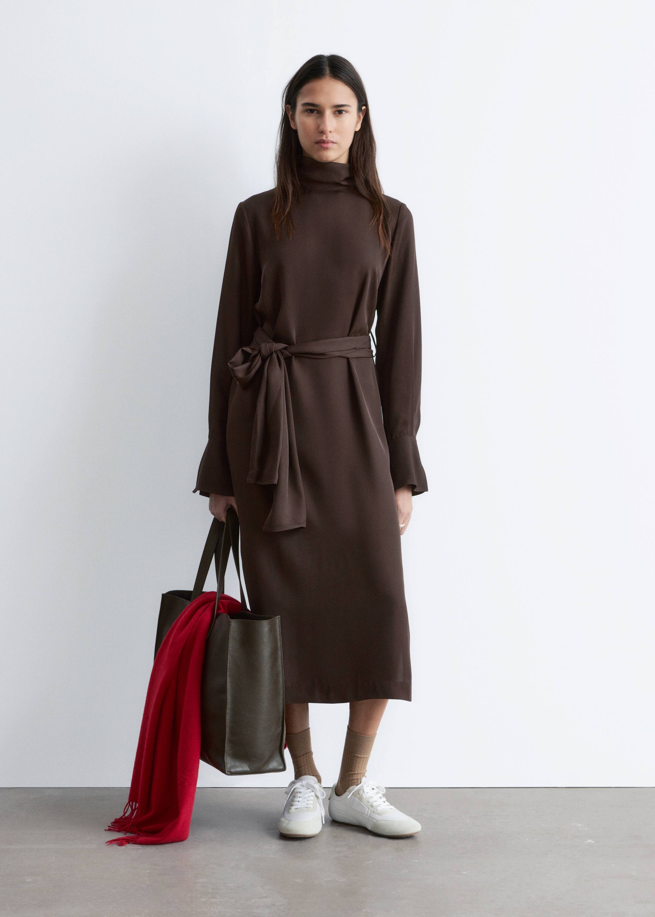 Vezi imaginea mai mare: Modelul poartă o rochie midi maro închis. Rochia are un decolteu înalt, mâneci lungi, ușor evazate, cu manșete cu nasturi și este strânsă în talie cu o curea din același material, căzând până la jumătatea gambei. Modelul ține, de asemenea, o geantă de mână închisă la culoare, care conține o eșarfă roșie.