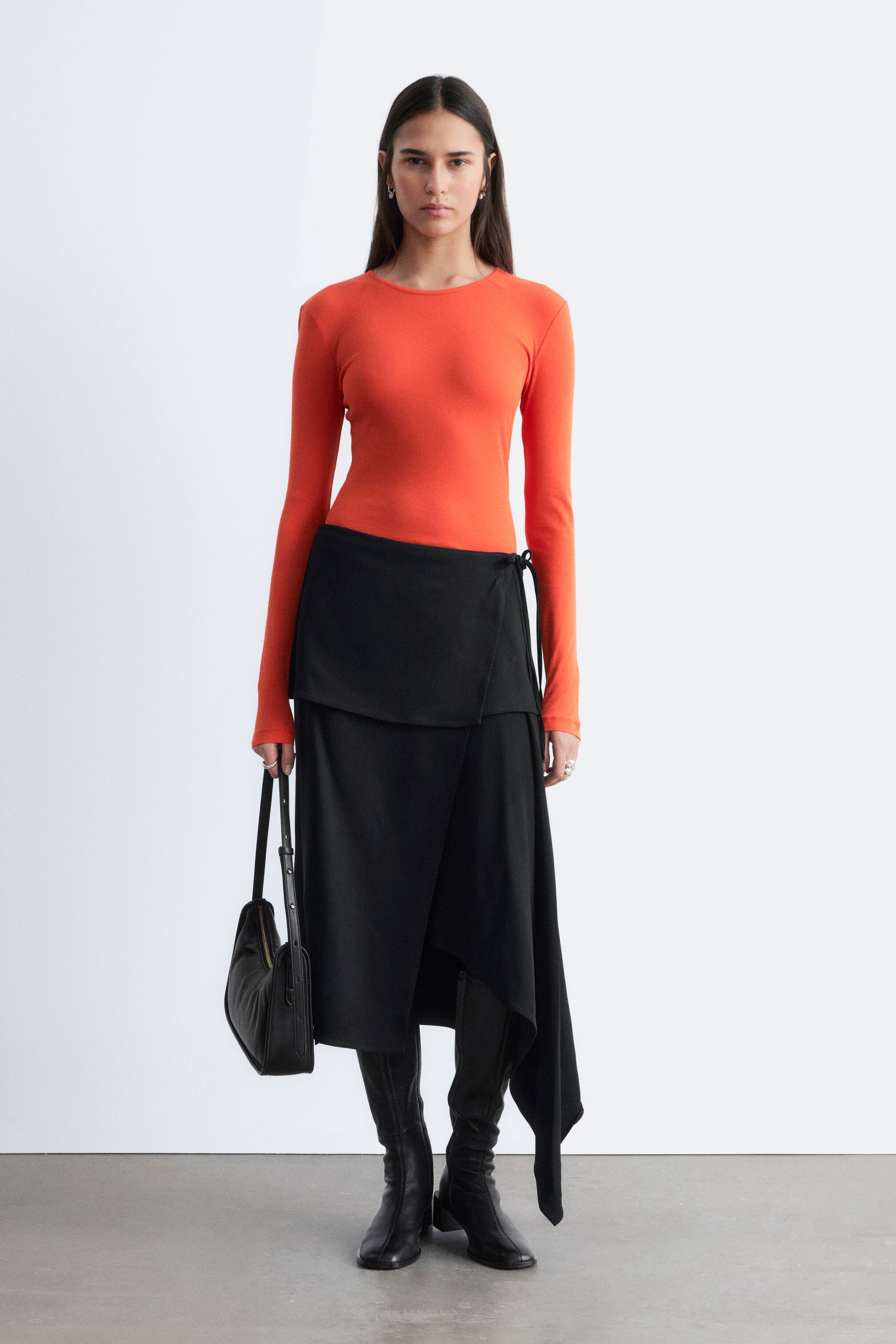Преглед на по-голямо изображение: Моделът носи черна пола с увиване, която се отличава с асиметричен силует с изчистен преден увивен дизайн и драпирано парче, падащо на една страна. Регулируеми връзки закрепват полата на талията, стилизирана върху оранжев топ с дълги ръкави и черни ботуши.