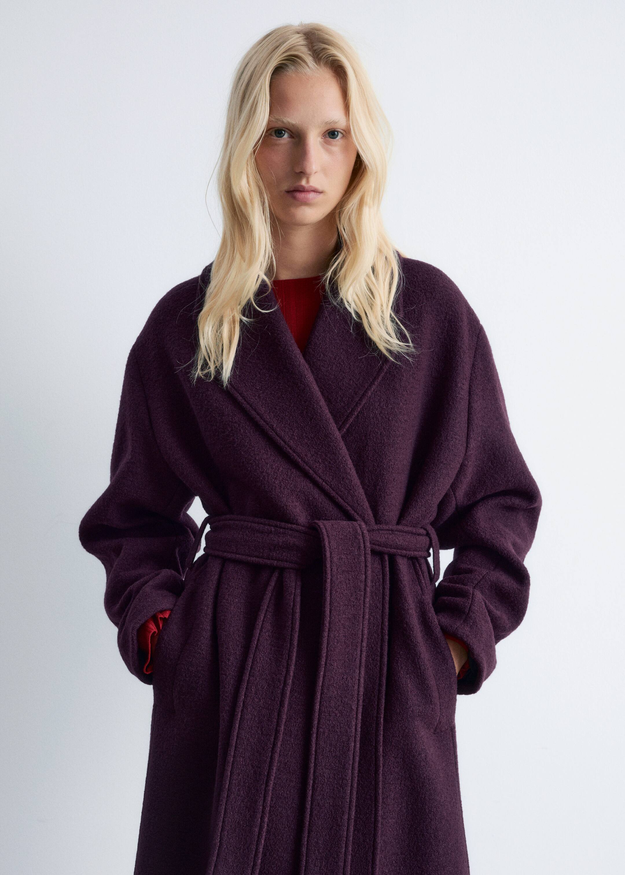 Agrandir l'image: Un manteau enveloppant texturé de couleur violet foncé est présenté avec de larges revers crantés et une ceinture à nouer à la taille par-dessus un vêtement rouge, affichant une coupe décontractée avec les mains reposant dans ses poches latérales.