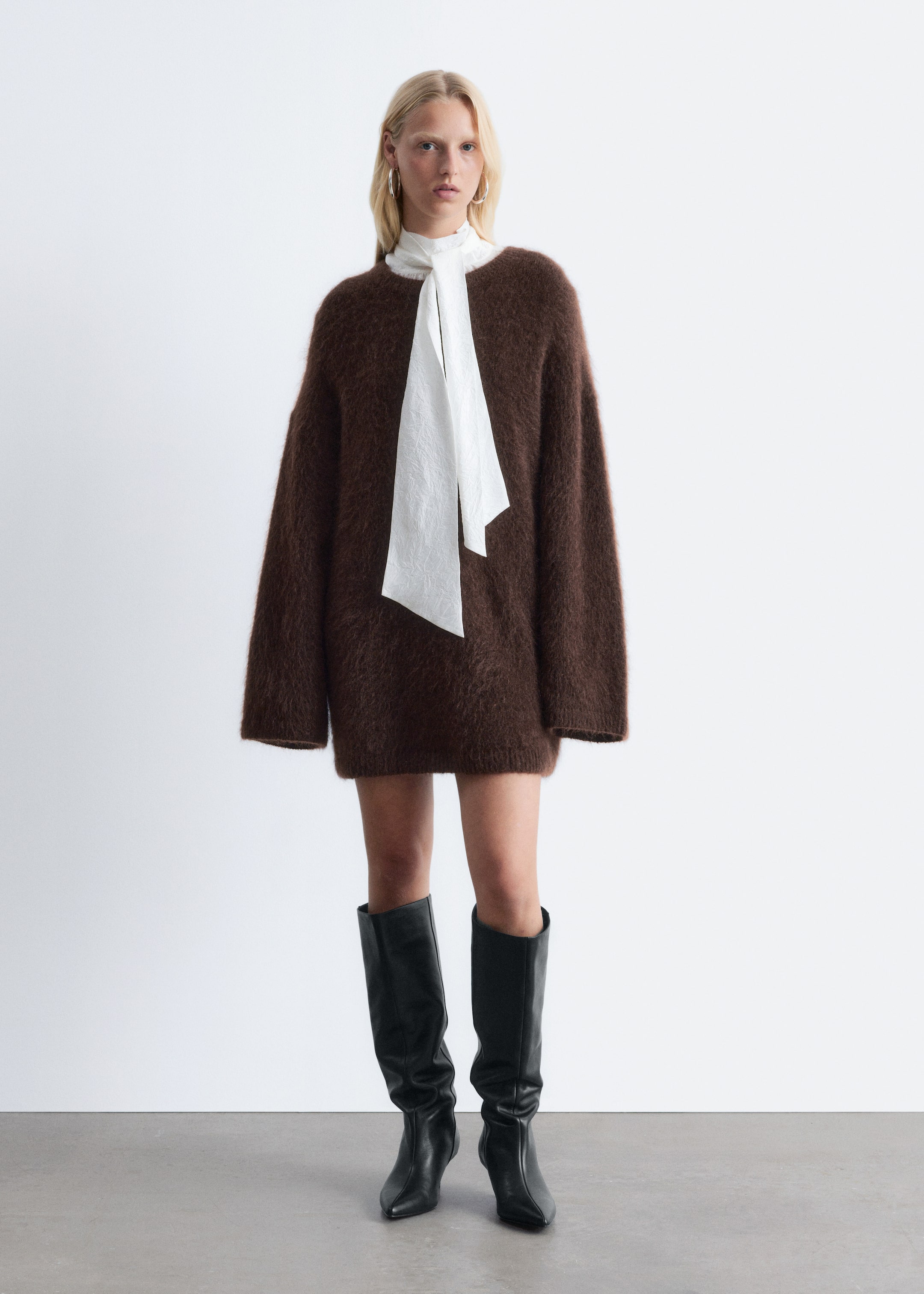 Visualizza immagine più grande: Un abito maglione oversize marrone e peloso viene indossato, rivolto in avanti, con maniche lunghe e larghe e un orlo che si ferma sopra il ginocchio. Una sciarpa bianca con texture viene stratificata sotto il collo rotondo, con le estremità che pendono davanti, e stivali neri alti fino al ginocchio vengono indossati con l'abito.