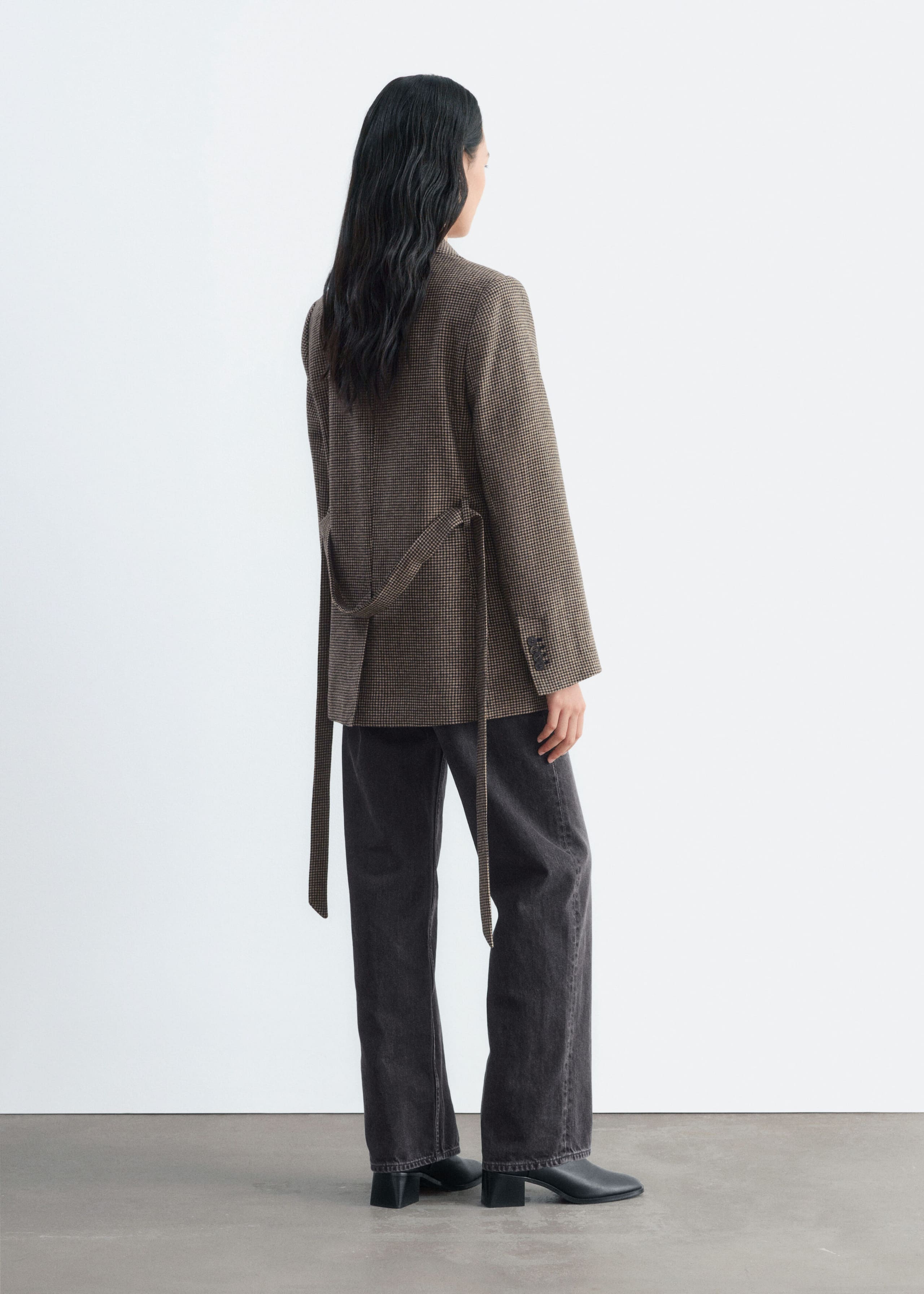 A brown houndstooth wool-blend blazer is being worn by the model, shown from a rear three-quarter angle. The blazer features a matching fabric tie belt hanging loosely from the waist and is paired with dark grey jeans and black boots.