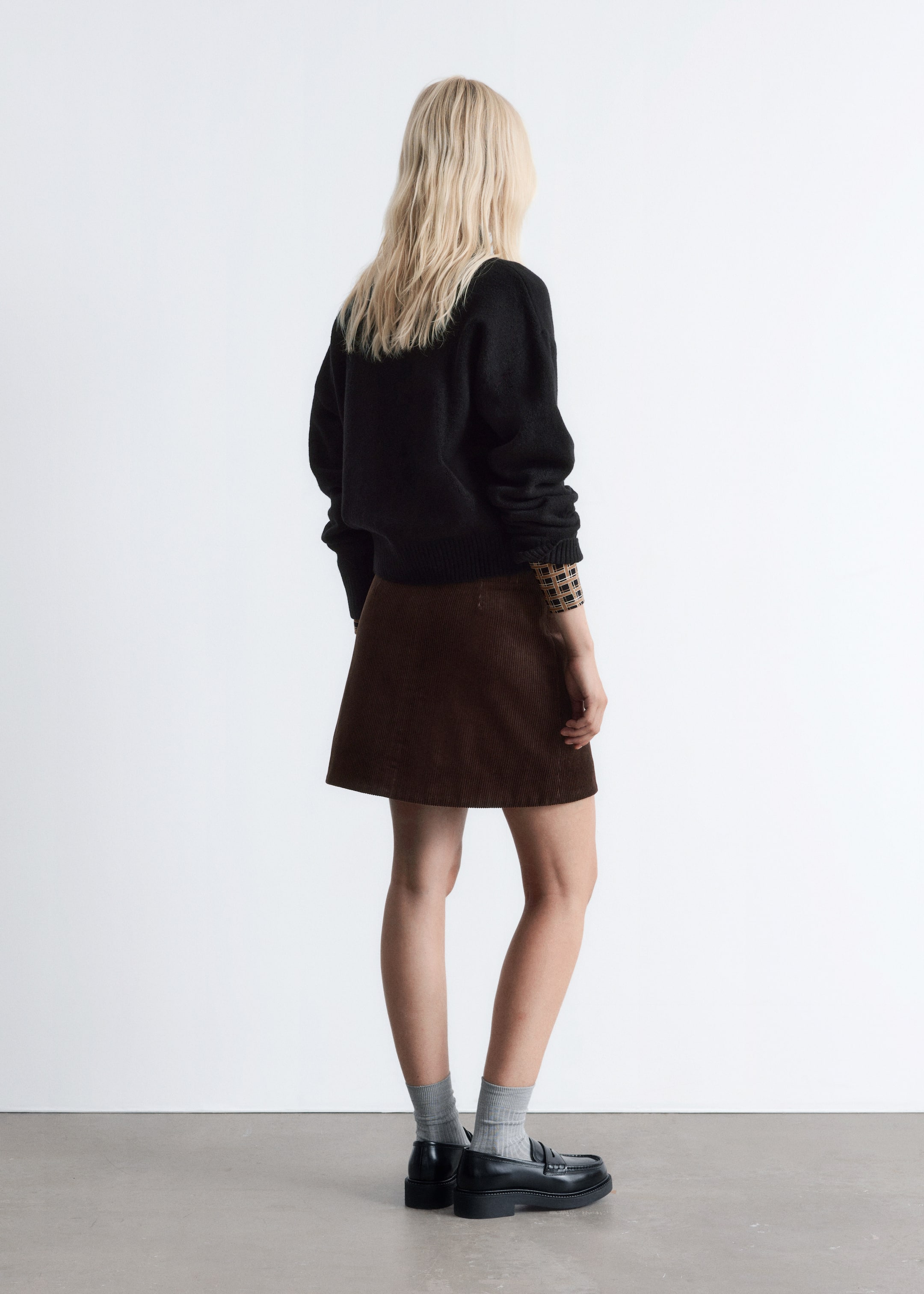 Größeres Bild anzeigen: Eine Person präsentiert ein Outfit aus einem Blickwinkel von hinten rechts, das einen schwarzen Pullover mit geripptem Saum und Bündchen zeigt, der über einem langärmeligen karierten Oberteil getragen wird. Sie tragen auch einen braunen Cord-A-Linien-Minirock, kombiniert mit grauen gerippten Socken und schwarzen Loafern mit dicken Sohlen.