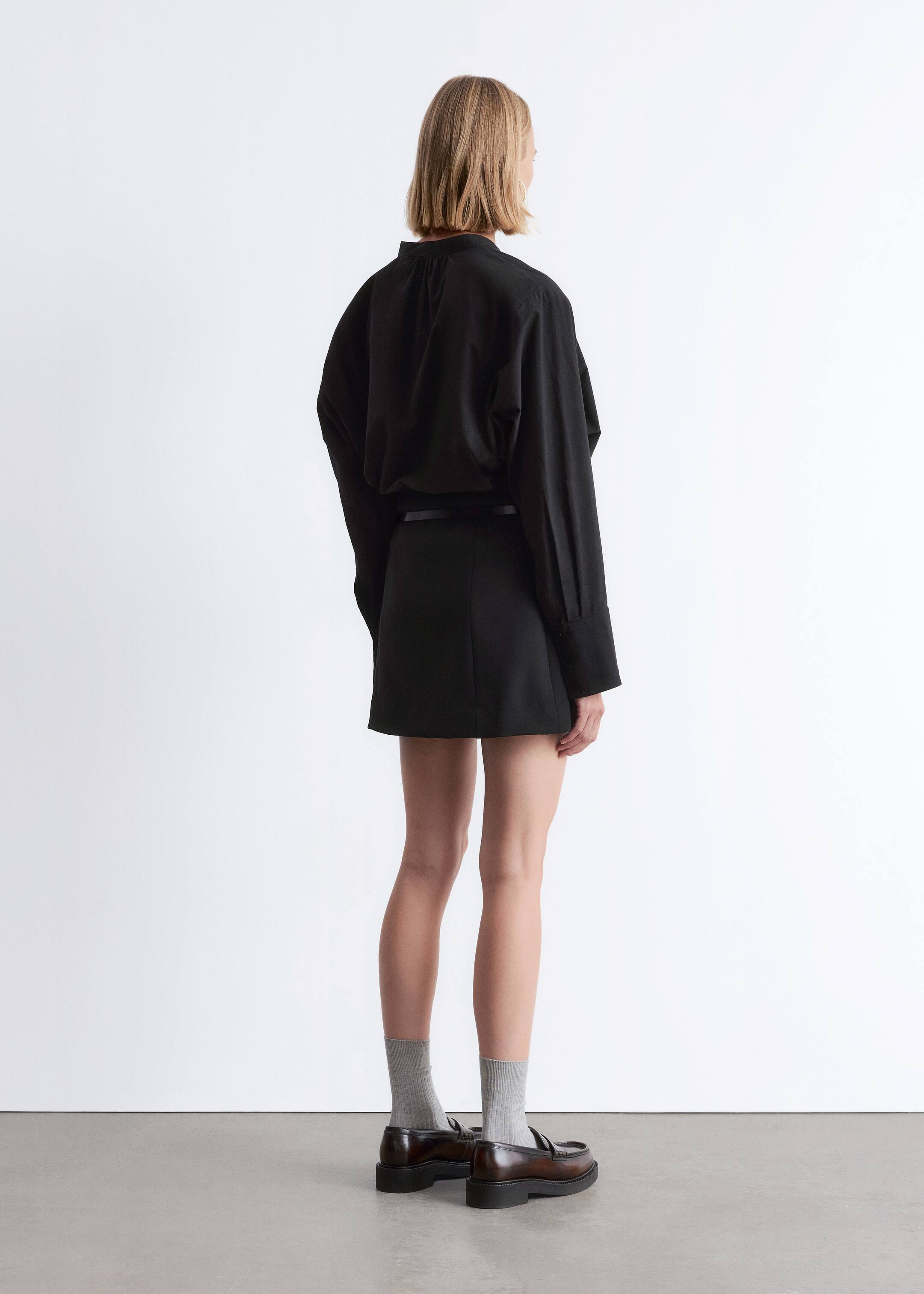 View larger image: A black oversized shirt, viewed from the back, is being worn with a black straight-cut mini skirt. The shirt features a gathered high neck and wide cuffs, and is being tucked into the skirt's fitted waist, creating a bloused effect over the skirt.