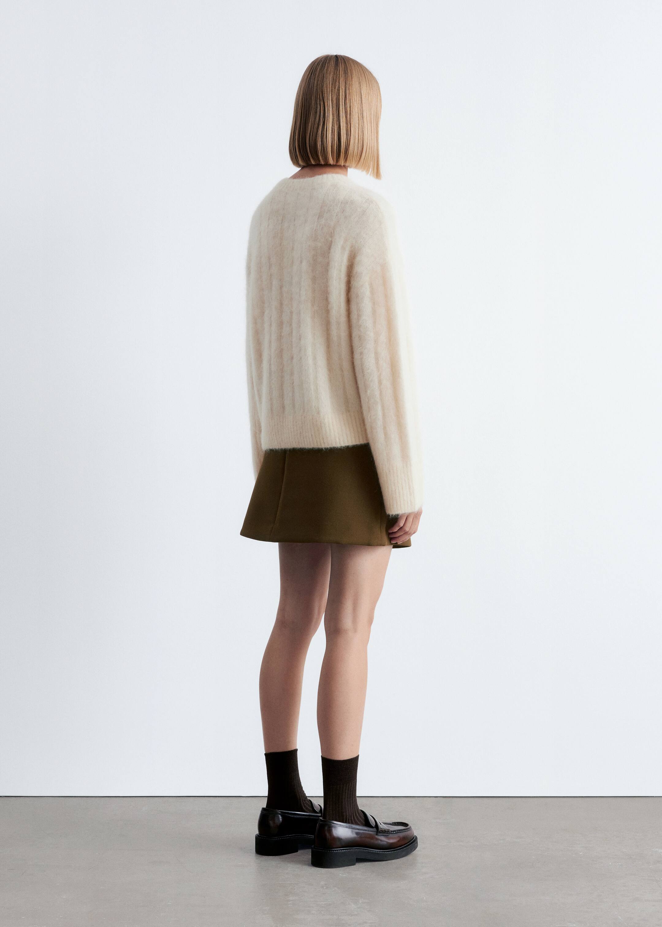 Se større billede: En cremefarvet, fuzzy ribstrikket sweater vises fra bagsiden og delvis sidevisning, med lange, afslappede ærmer og en lige kant. Den er parret med en olivengrøn A-line mininederdel, mørkebrune ribstrikkede sokker og chunky brune loafers.