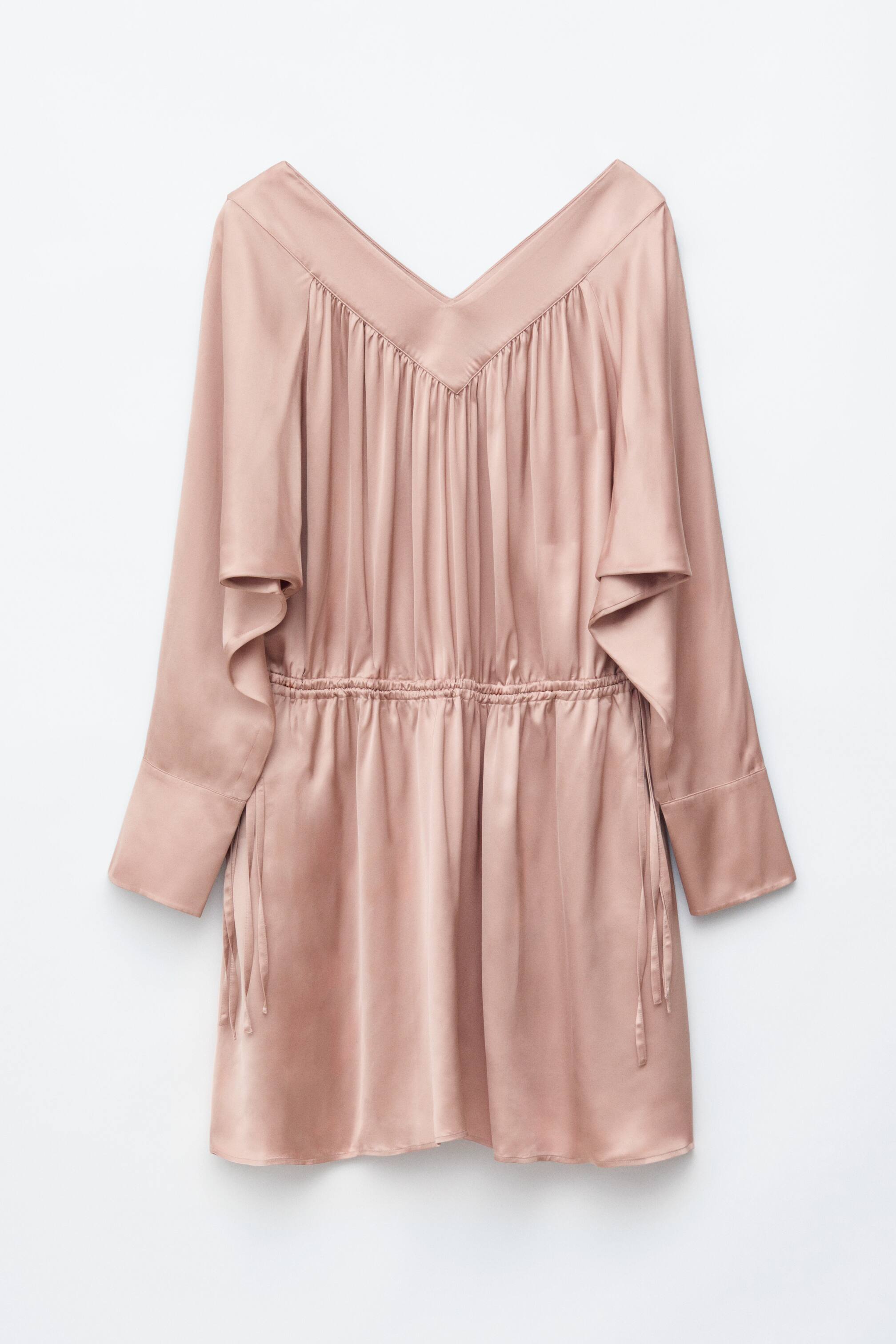 Agrandir l'image: Une robe en satin rose clair présente son devant, avec un profond décolleté en V et de nombreux plis sur le buste. Elle est dotée de manches longues avec de larges poignets et d'une taille cintrée avec cordon de serrage, dont les liens pendent sur les côtés.