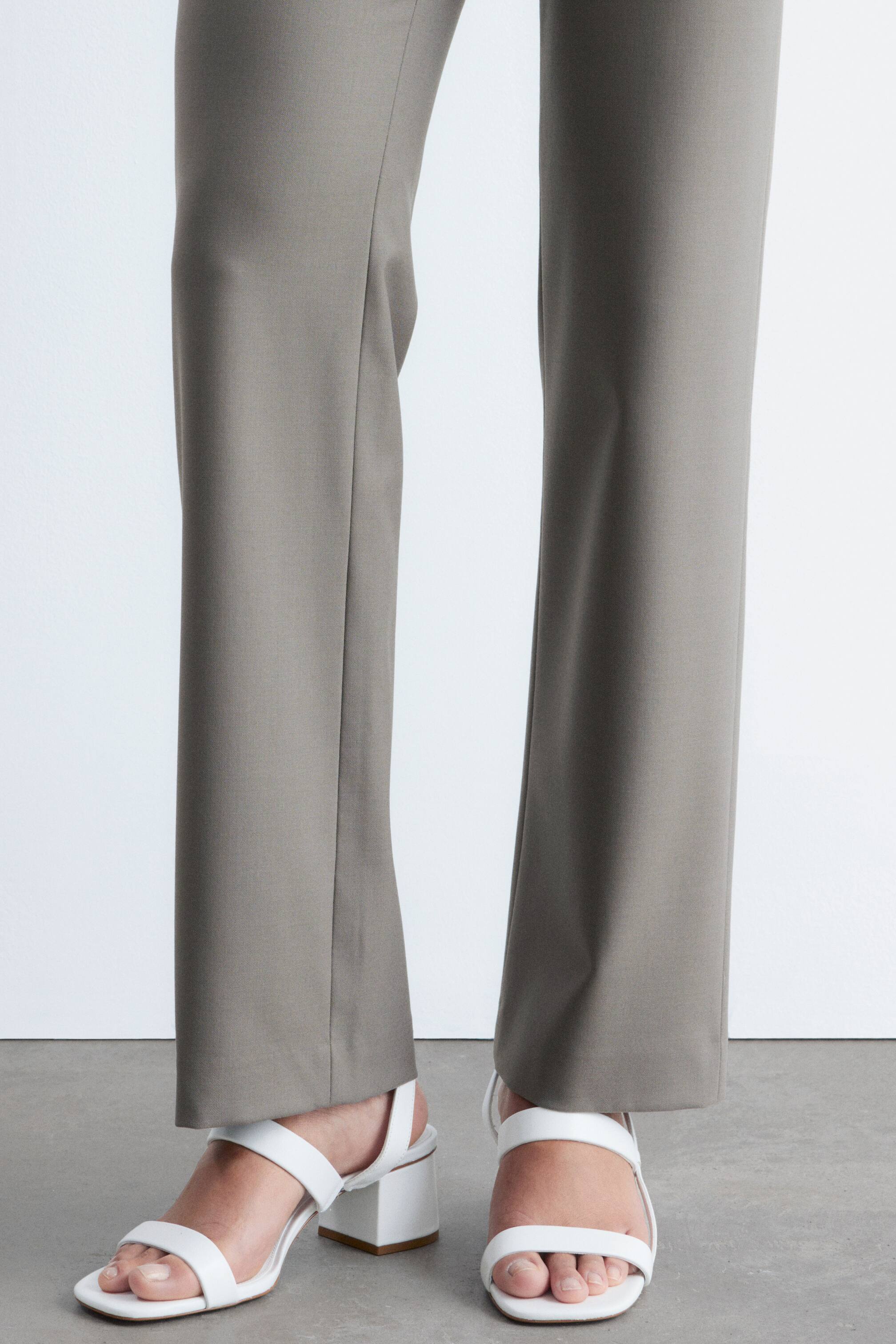 Visa större bild: Taupe byxor bärs, med en rak passform och ett slätt tyg. De faller rakt ner och når precis ovanför vita blockklackade sandaler.