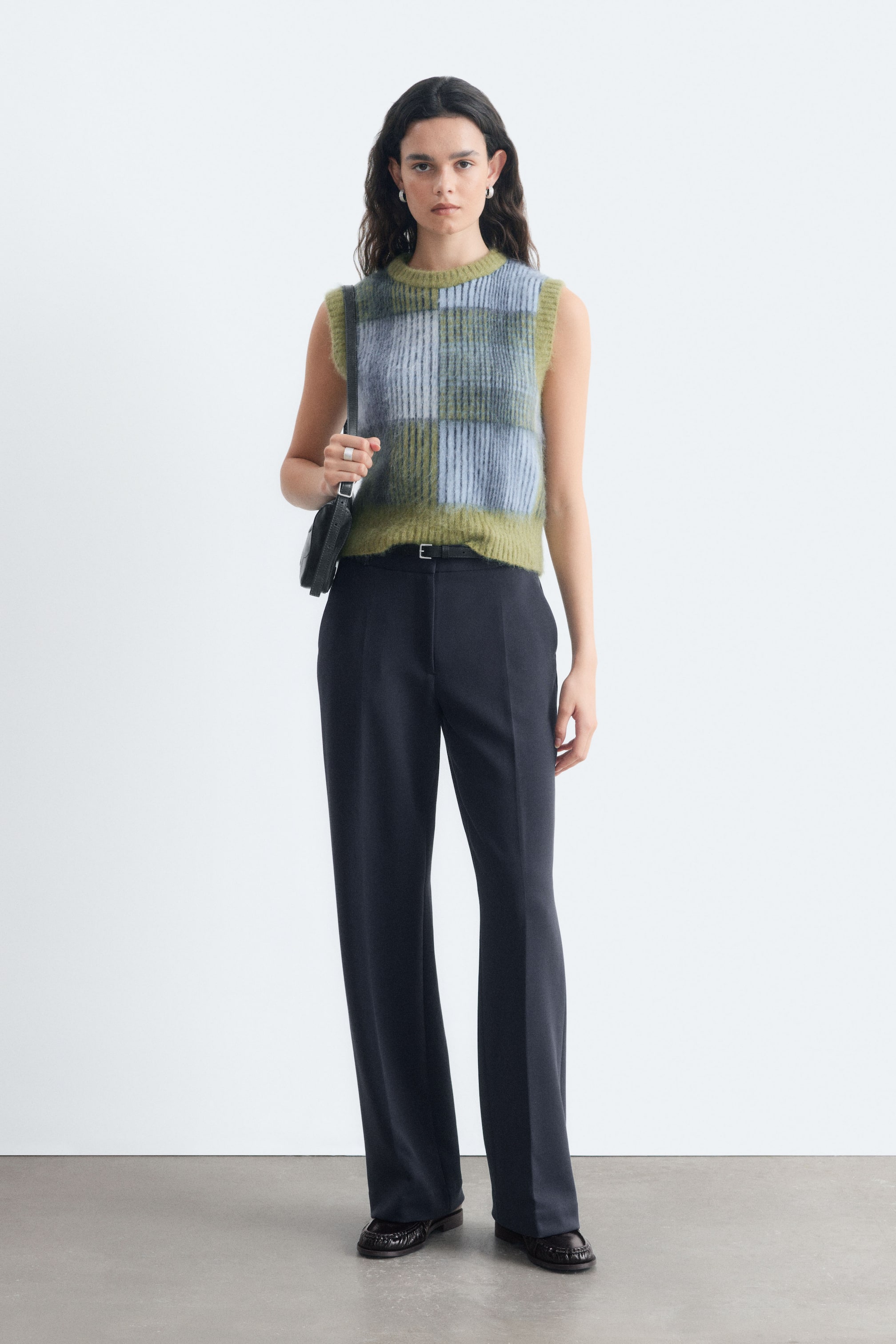 View larger image: Dark straight-leg trousers feature pressed creases, a waistband with belt loops, and slanted side pockets. The model is wearing the trousers, styled with a slim black belt.