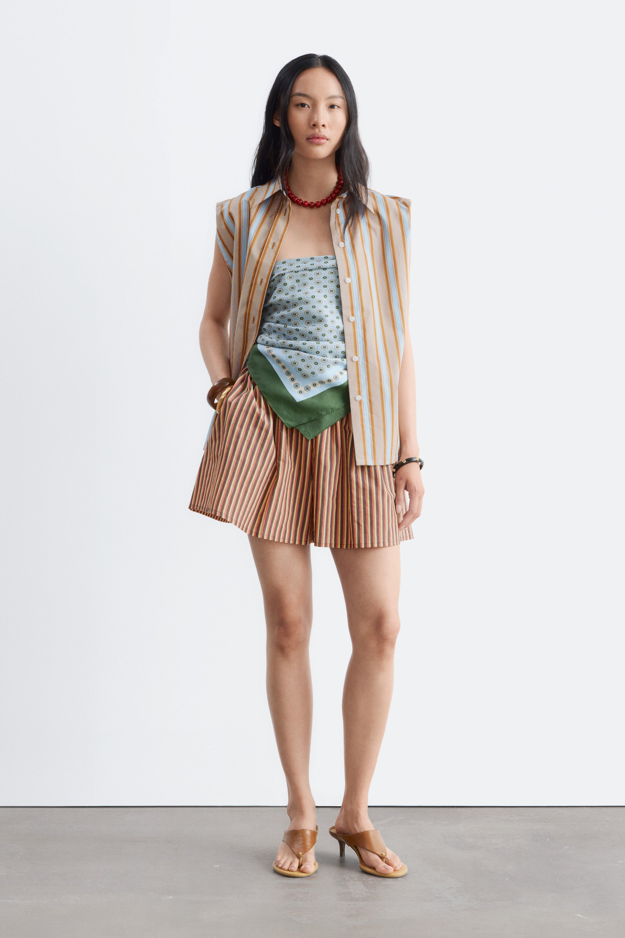 Ver imagem maior: O produto está a ser apresentado de frente, com uma camisa sem mangas com colarinho e riscas verticais em bege e azul claro, usada aberta sobre um top bandeau azul claro com padrão circular e uma borda verde escura. Uns calções plissados com riscas verticais em bege e castanho-avermelhado são usados, juntamente com sandálias de dedo em couro castanho com um pequeno salto.