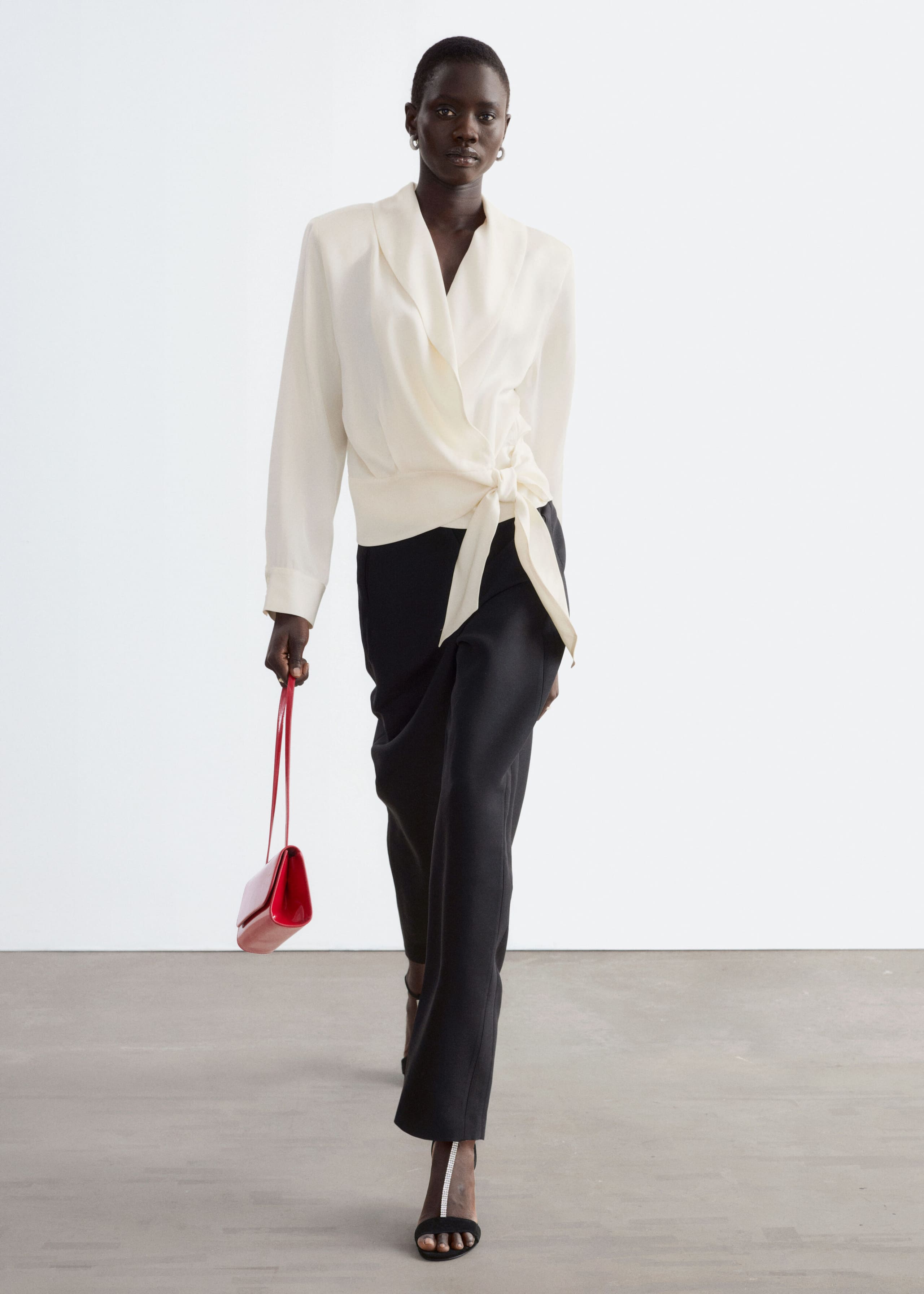 La modella indossa una blusa avvolgente color crema con collo sciallato e vita con chiusura laterale. La blusa è abbinata a pantaloni neri e la modella porta una piccola borsa a mano rossa.