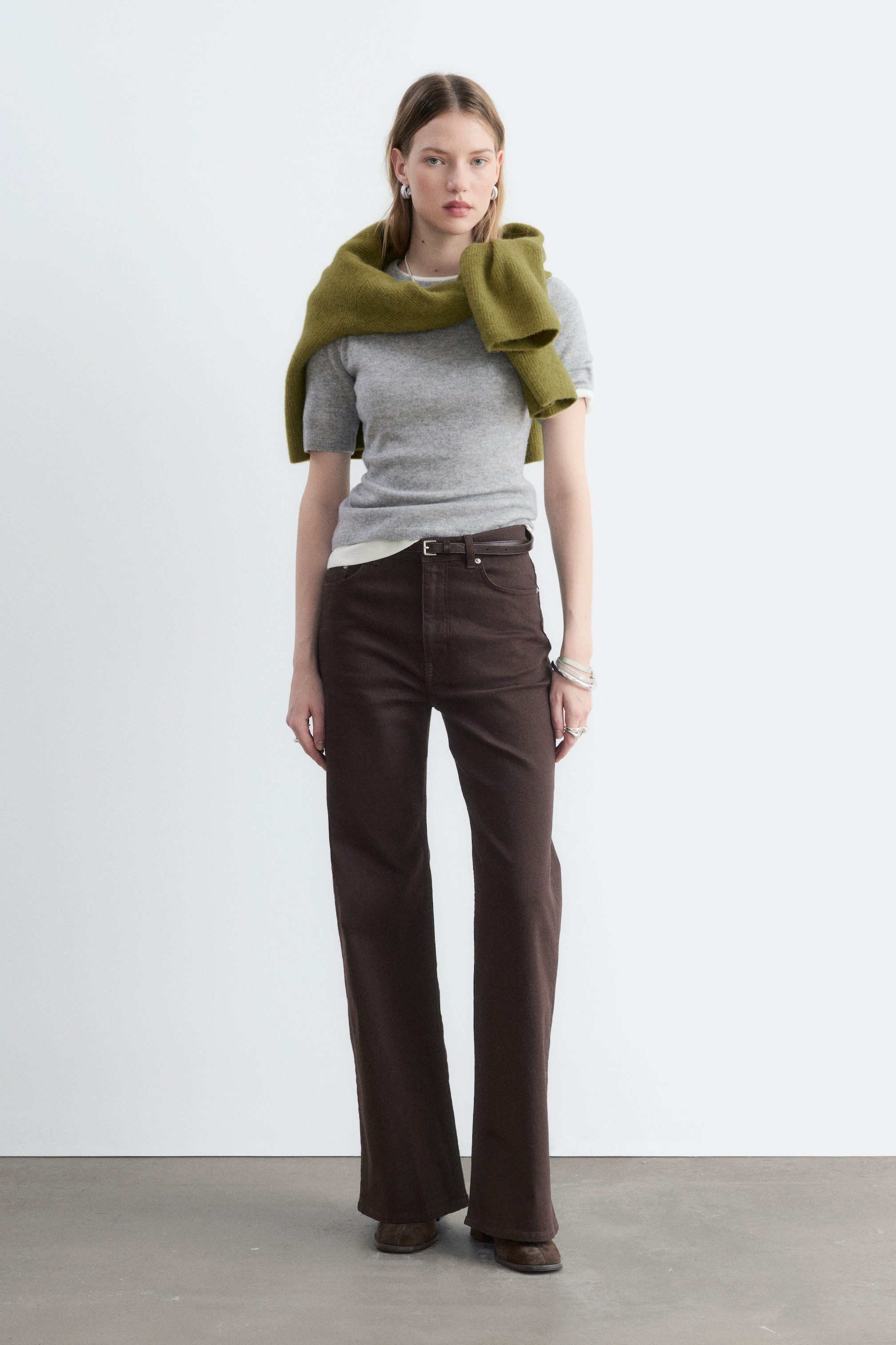 Visualizza immagine più grande: Jeans marroni scuri a vita alta, dalla vestibilità regular e con gambe lunghe e ampie, indossati dalla modella. I jeans sono abbinati a una sottile cintura marrone, un top grigio a maniche corte e un maglione verde appoggiato sulle spalle.