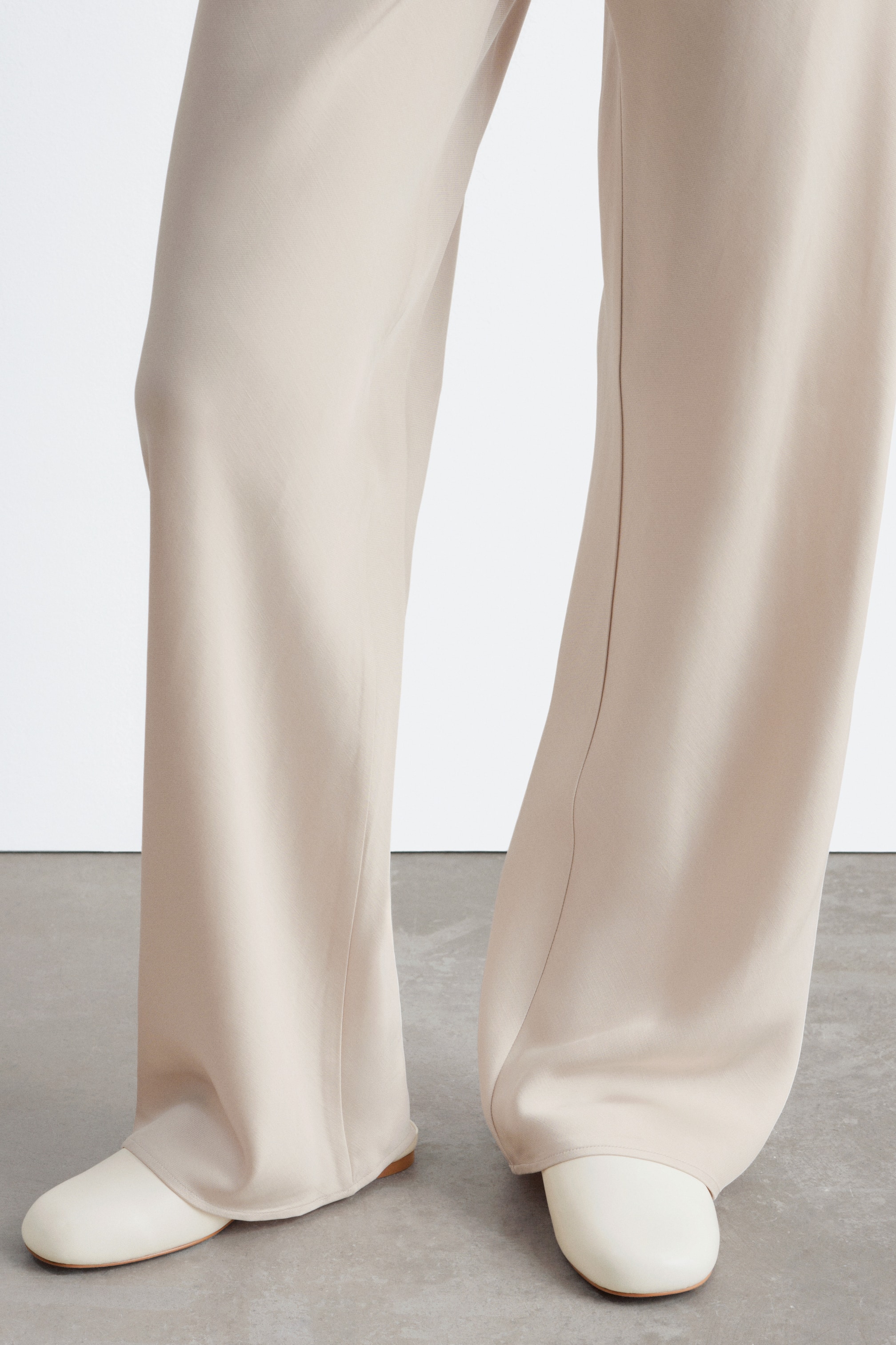 Visualizza immagine più grande: Pantaloni beige a gamba larga che si adagiano su scarpe piatte color crema, con ogni gamba del pantalone caratterizzata da una piega frontale stirata.