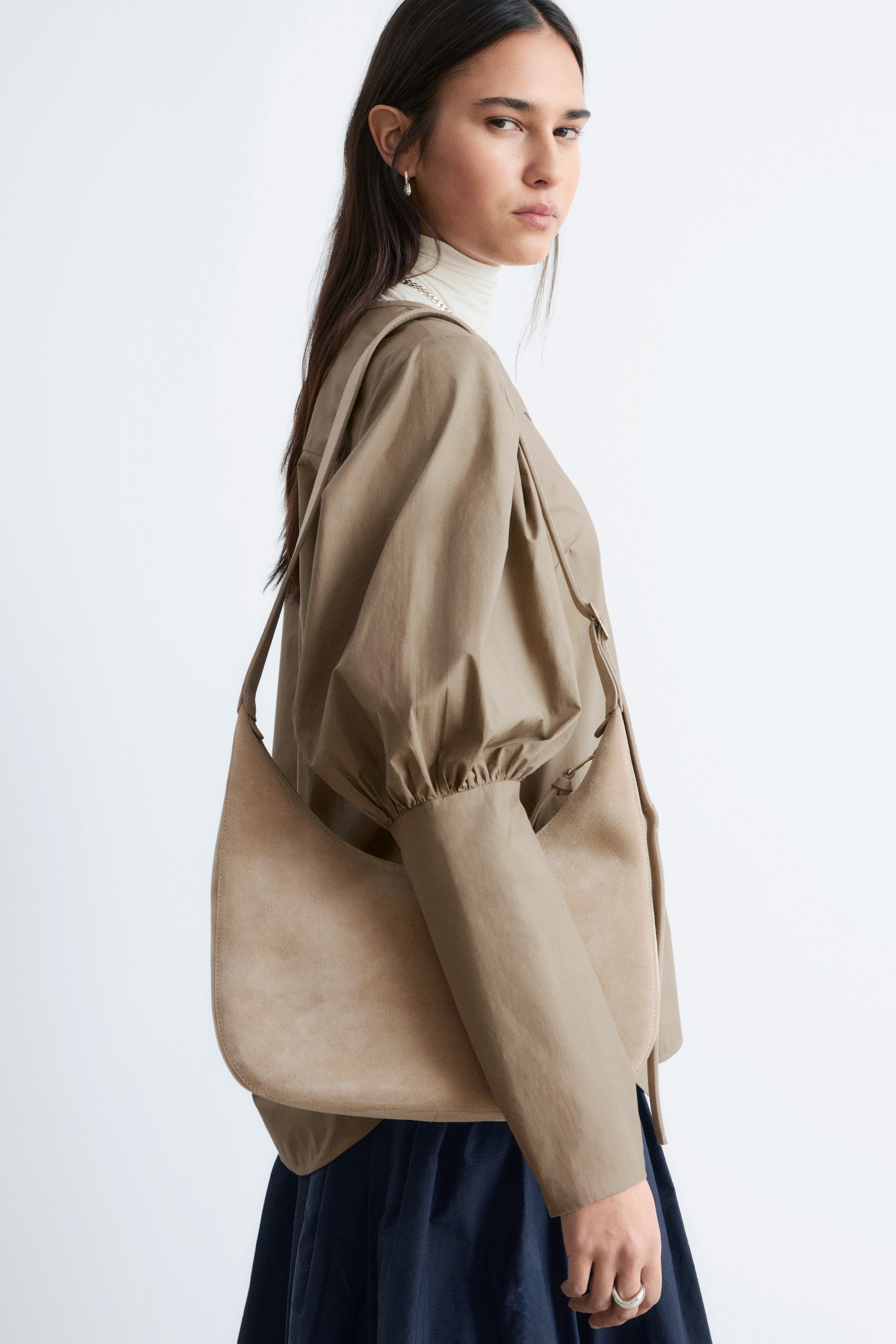 Ver imagen más grande: Un bolso de hombro en forma de media luna de color marrón claro, que parece estar hecho de ante, se lleva en el hombro derecho. Su cuerpo suave se curva bajo el brazo, mostrando la textura lisa y la silueta redondeada del bolso.