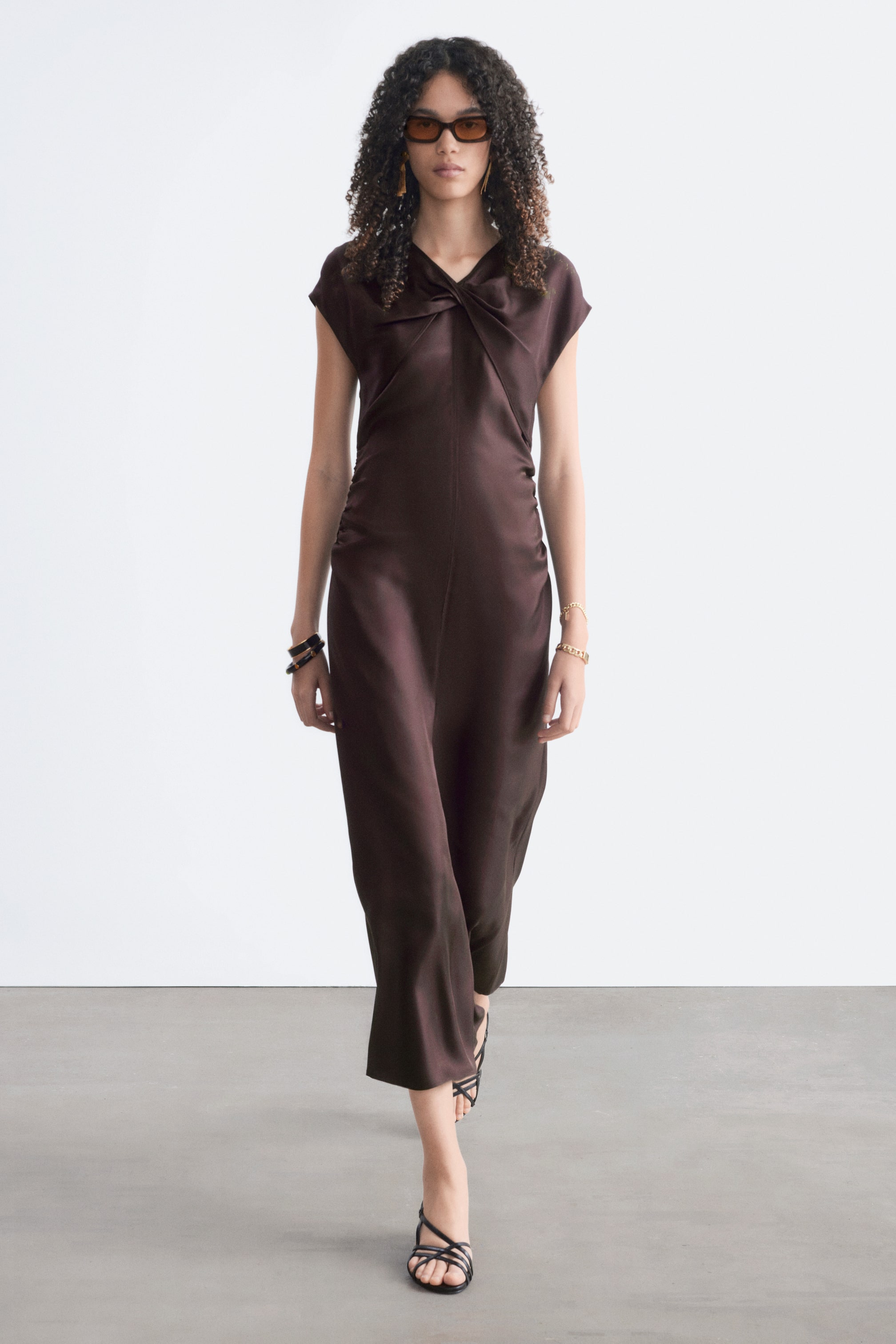 View larger image: The dark brown midi dress features a V-neckline with a twisted knot detail at the bust and short cap sleeves. It presents a flowing silhouette with gentle side ruching and a subtle sheen.