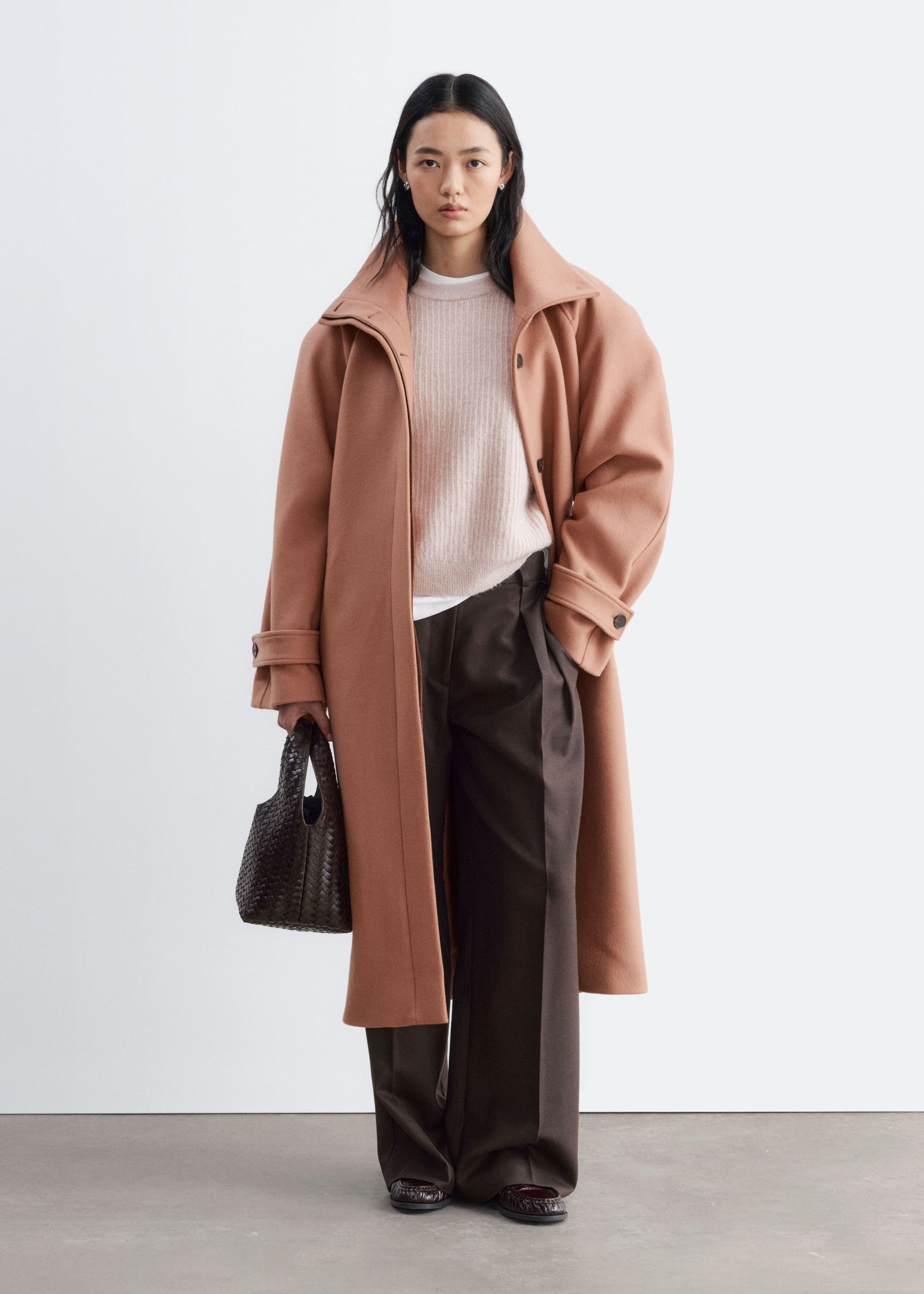 A peach-coloured wool-blend coat featuring a wide collar, dropped shoulders and buttoned cuffs is worn open by the model. The coat is layered over a light pink knit jumper and wide-leg dark brown trousers.