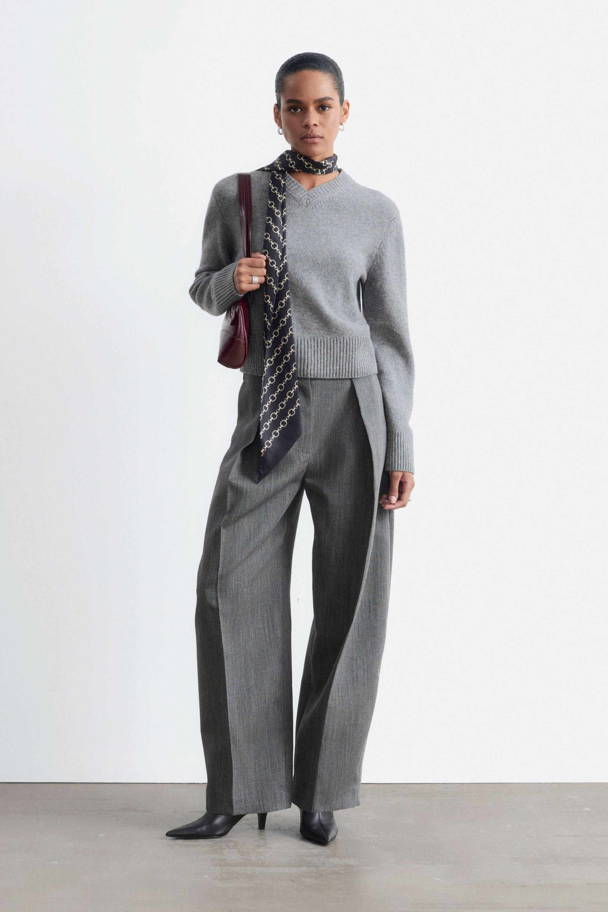 Visualizza l'immagine ingrandita: Un maglione grigio lavorato a maglia con scollo a V a costine, orlo ampio a costine e polsini a costine è indossato dalla modella. Il maglione è abbinato a pantaloni grigi a gamba larga, una sciarpa a fantasia drappeggiata intorno al collo e una borsa a tracolla rosso scuro.
