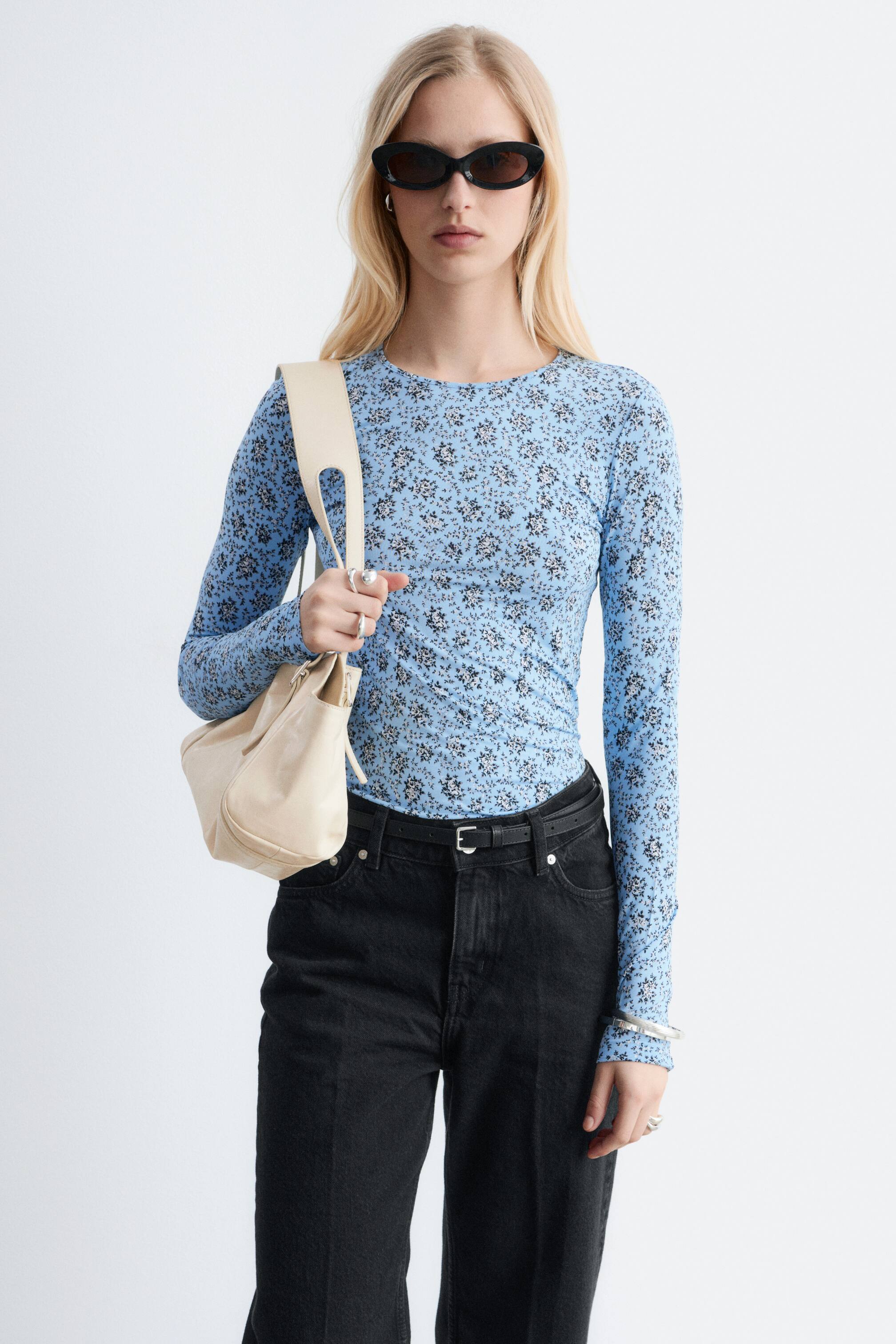 Ver imagem maior: A camisola de manga comprida apresenta uma base azul clara com um padrão floral disperso em preto e branco. É desenhada com uma gola redonda e mangas compridas, exibindo uma silhueta ajustada e sendo usada por dentro de jeans pretos.