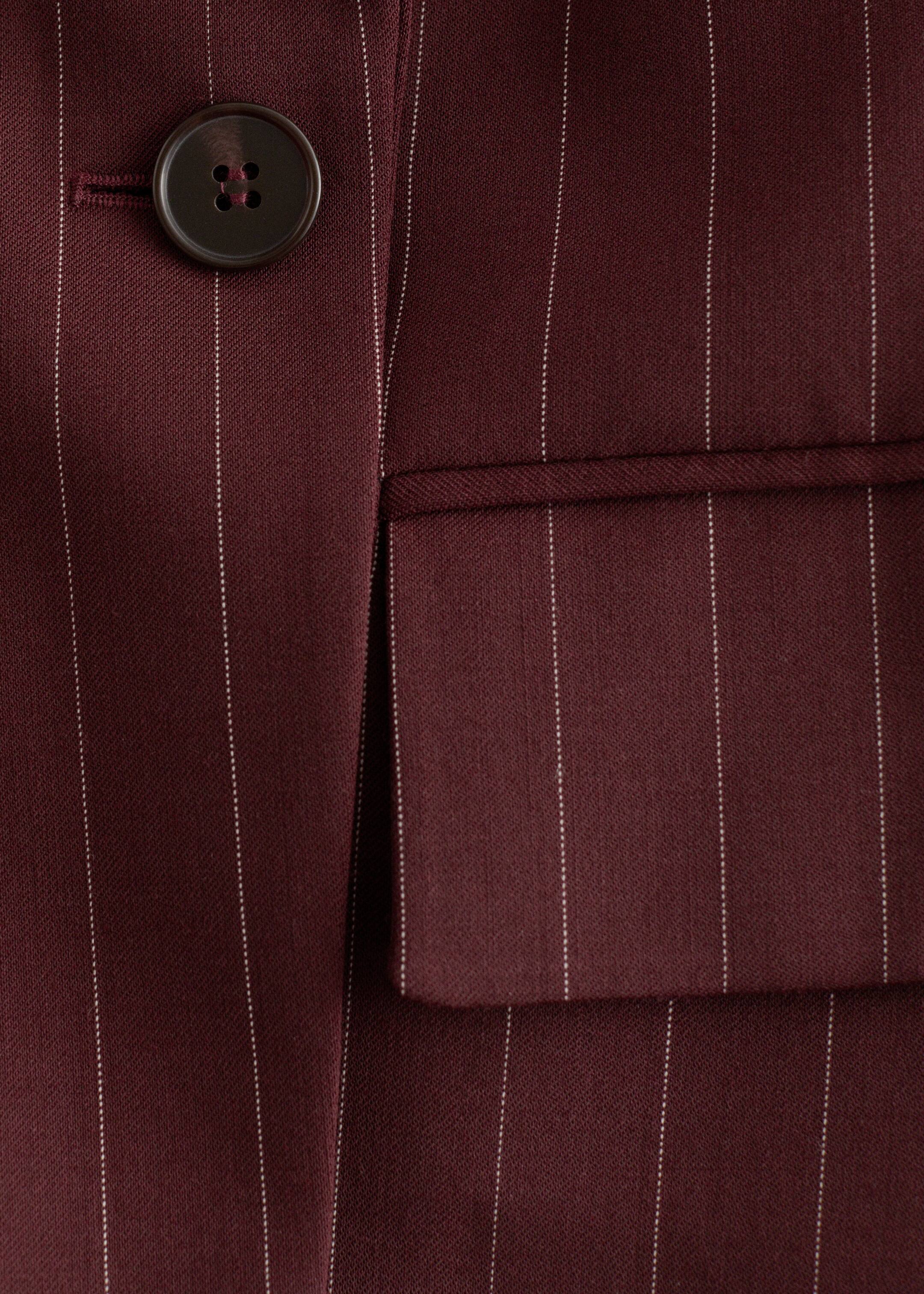 Agrandir l'image: Un blazer bordeaux à fines rayures est présenté en gros plan, avec de fines rayures verticales blanches. Un bouton marron foncé à quatre trous est visible à gauche, et une poche à rabat apparaît à droite.