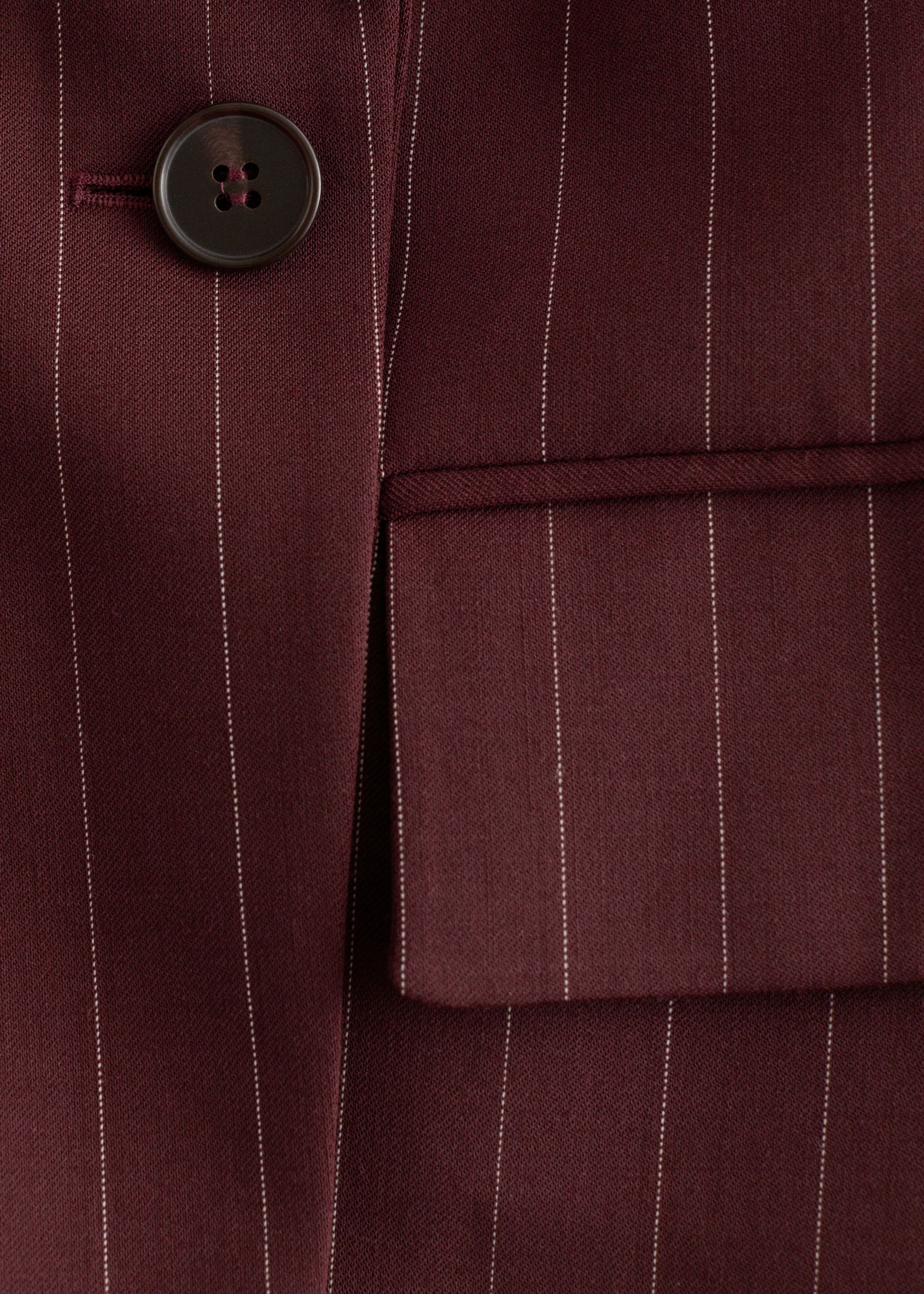 Grotere afbeelding bekijken: Een bordeauxrode blazer met krijtstreep wordt in close-up getoond, met fijne witte verticale krijtstrepen. Een donkerbruine knoop met vier gaten is zichtbaar aan de linkerkant, en een klepzak verschijnt aan de rechterkant.