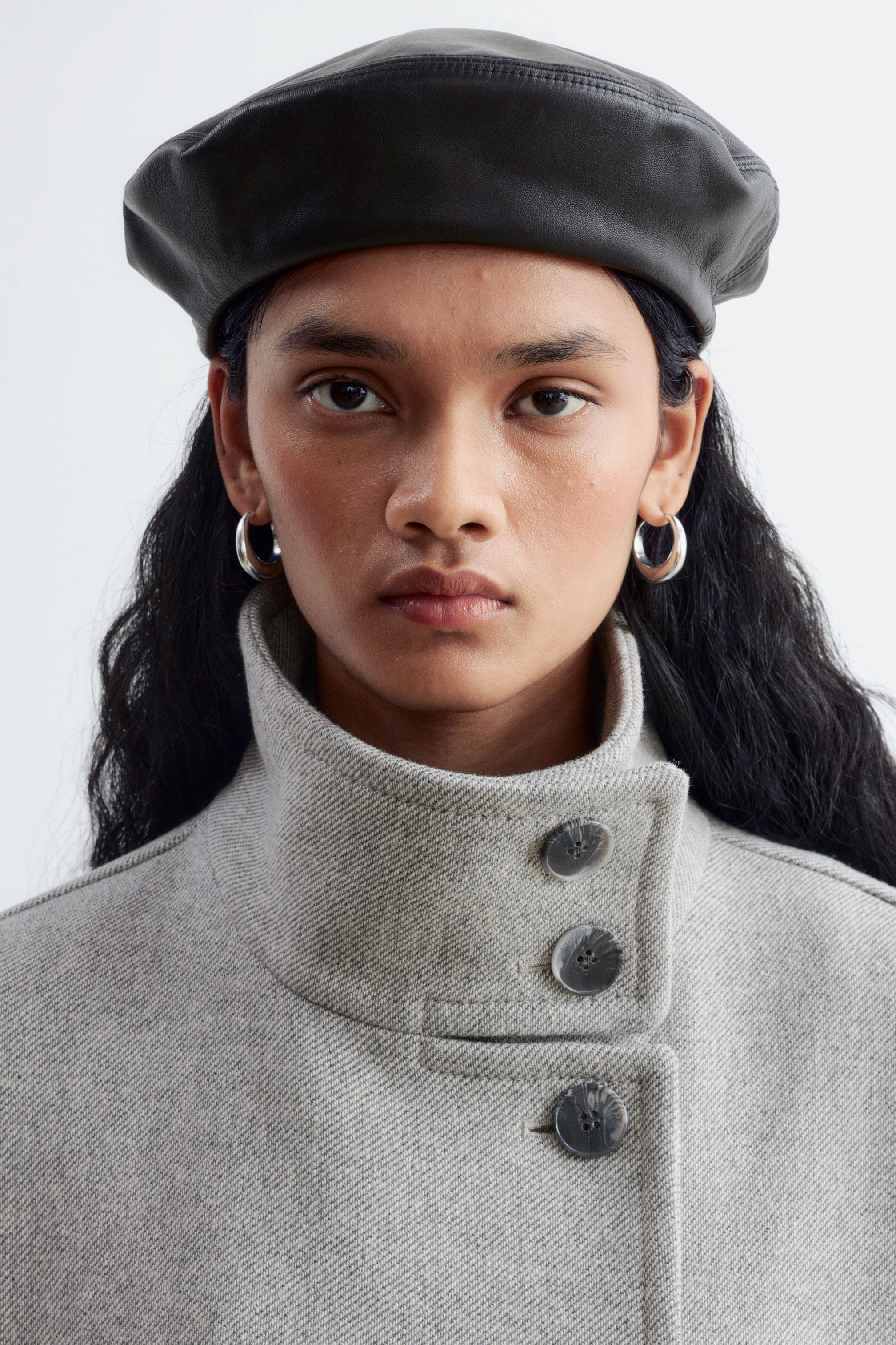 Grösseres Bild ansehen: Ein schwarzes Leder-Beret mit glatter, strukturierter Oberfläche wird auf dem Kopf des Models getragen. Das Beret ist zusammen mit einem grauen Mantel mit hohem Kragen und Knopfleiste gestylt.