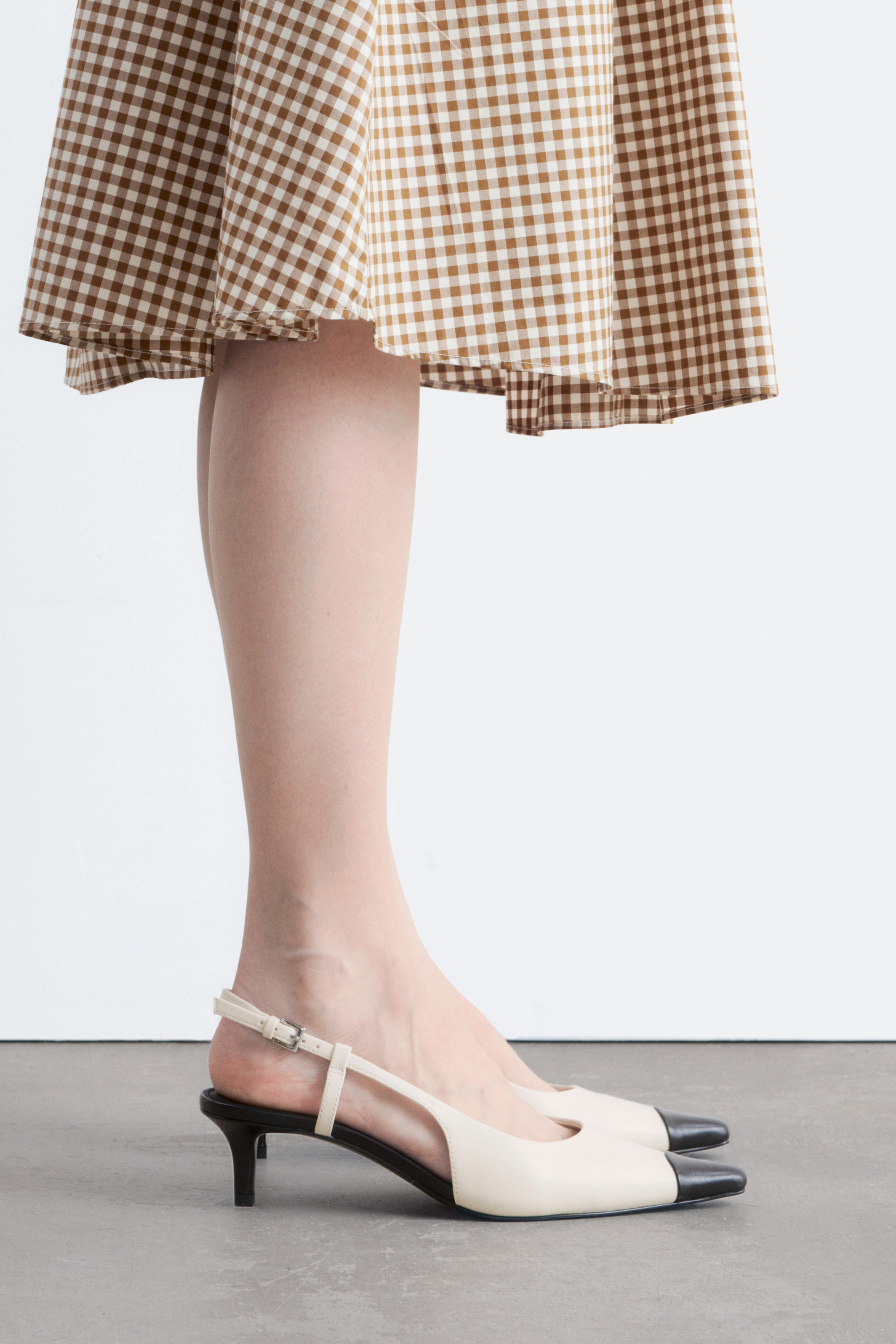 Ver imagen más grande: Los zapatos de tacón con tira trasera presentan un cuerpo en beige claro con una puntera en contraste de color negro. Están diseñados con un tacón bajo de color negro y una correa ajustable beige en la parte trasera, presentándose desde un perfil lateral.