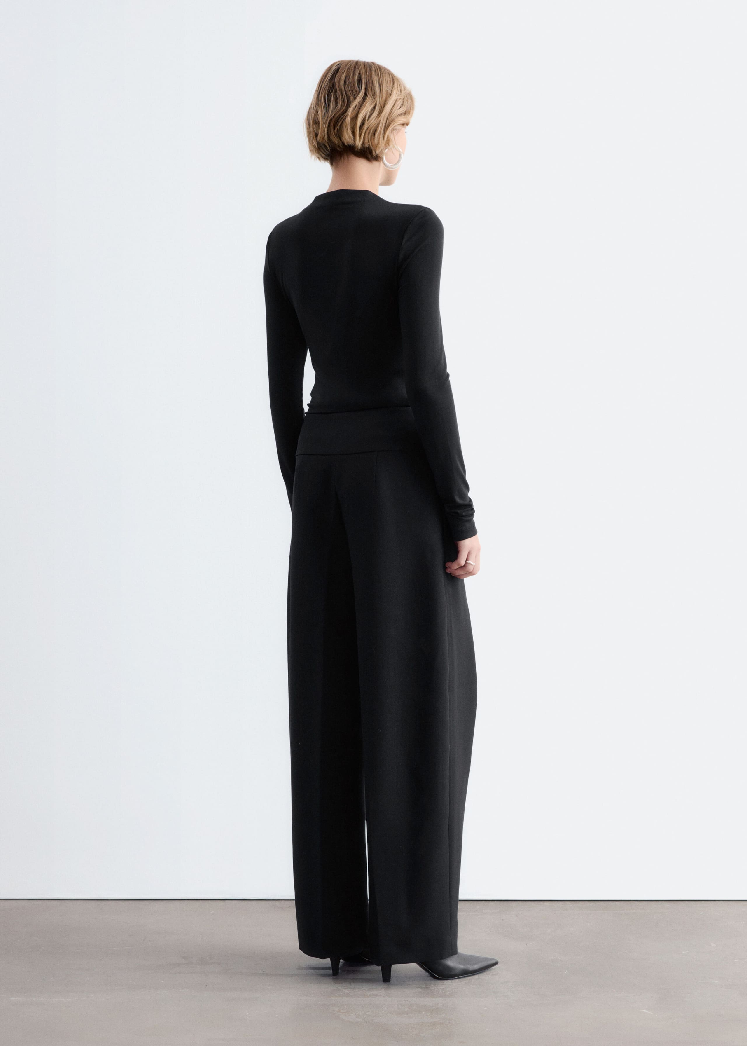 A black, close-fitting, long-sleeved top is being worn by the model, shown from the back. The top is styled with matching black wide-leg trousers and black pointed-toe heeled shoes.
