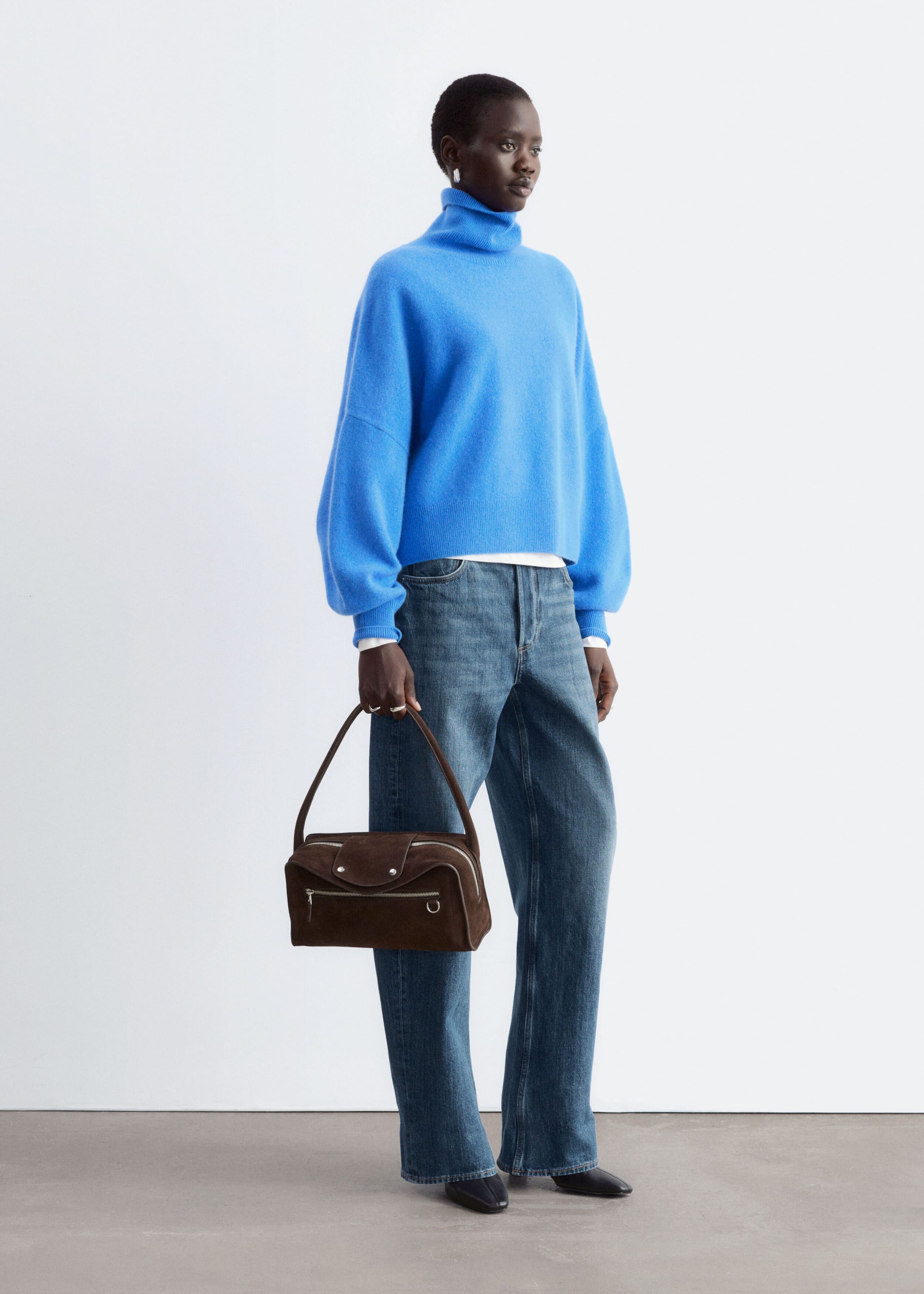 An oversized blue knitted jumper with a high folded roll neck and dropped shoulders is being worn by the model. The jumper is styled with wide-leg denim jeans, and the model is holding a brown suede handbag.