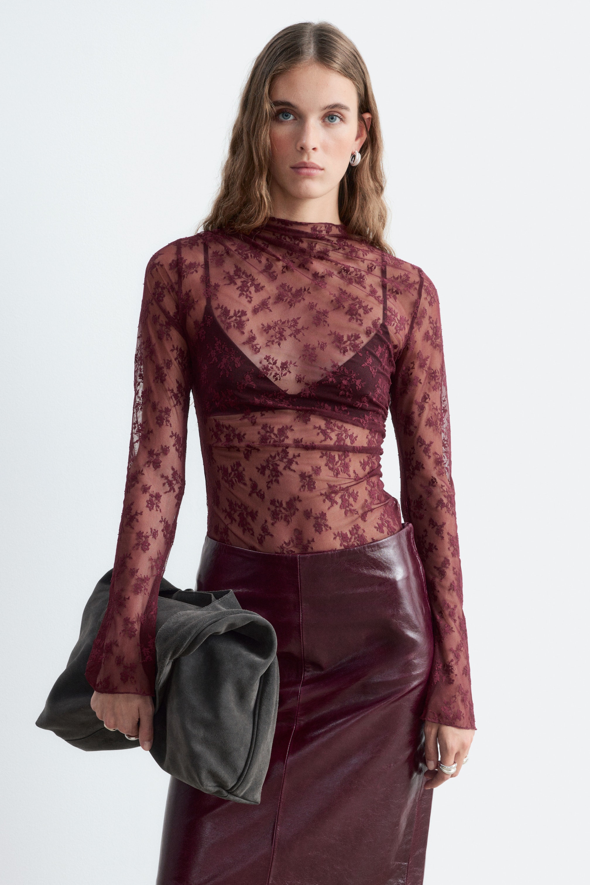 Agrandir l'image: Un haut en dentelle florale bordeaux transparente à col montant et manches longues évasées est porté par le mannequin par-dessus un soutien-gorge assorti. Le haut est rentré dans une jupe en cuir bordeaux, et le mannequin tient une grande pochette gris foncé.