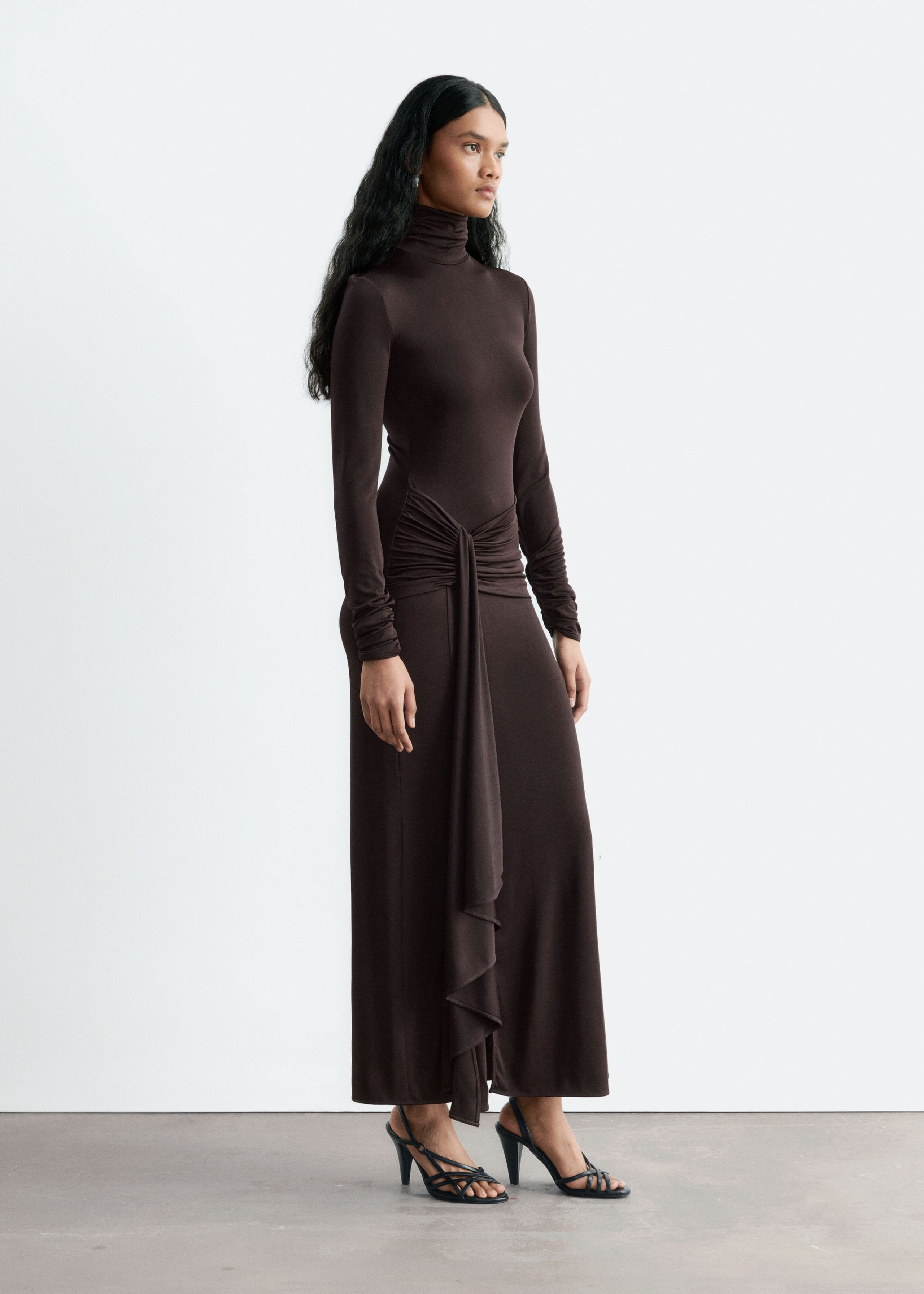 A dark brown, slim-fit jersey dress featuring a mock neck, long sleeves and a draped tie detail at the waist with a cascading fabric panel is worn by the model. The ankle-length dress is styled with black strappy high-heeled sandals.