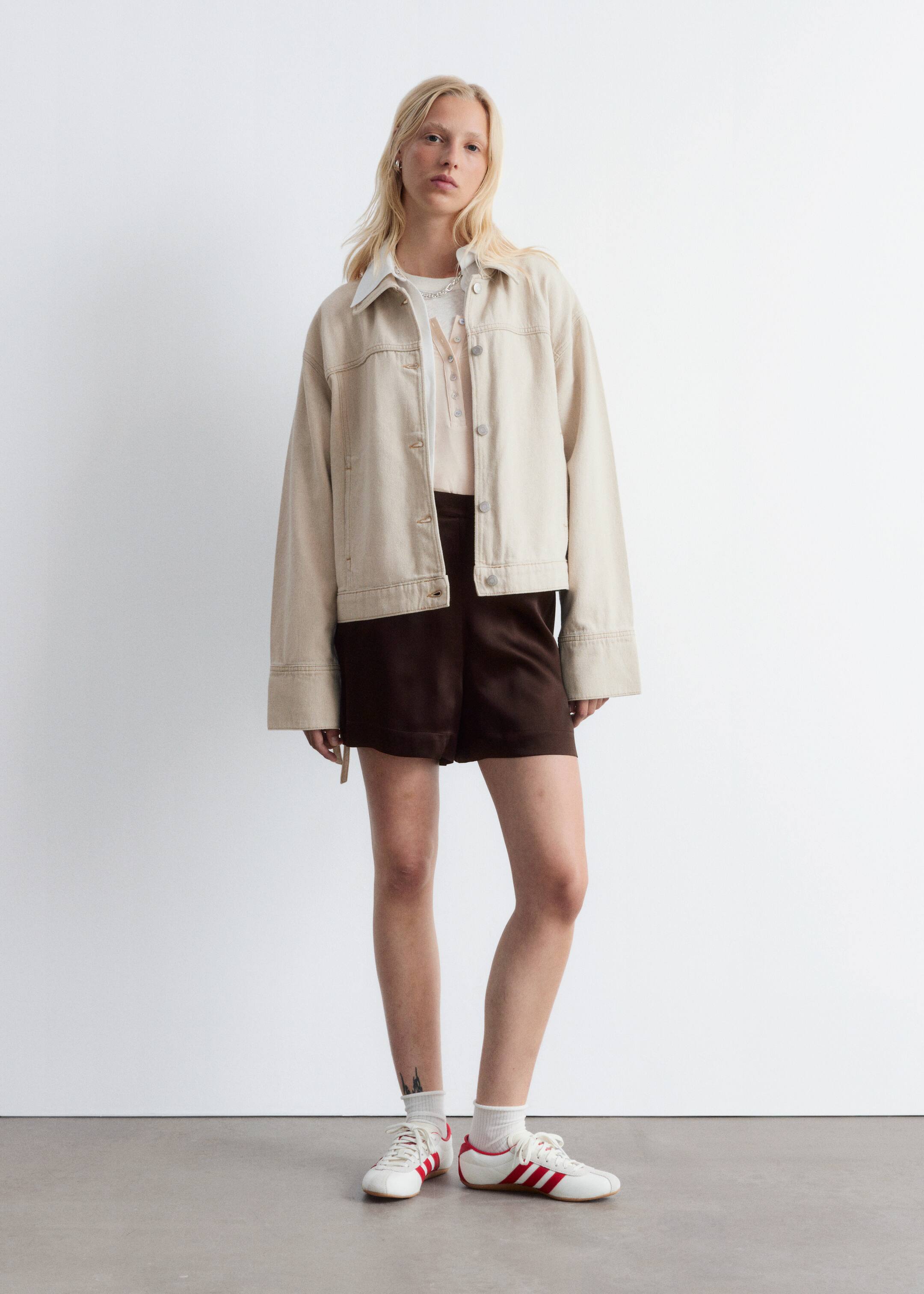 Agrandir l'image: Une veste de style denim de couleur beige clair est présentée, avec une coupe décontractée et oversize avec des épaules tombantes. Il est conçu avec un col pointu, une fermeture à boutons argentés sur le devant et deux poches poitrine à rabat boutonné, le tout accentué par des coutures contrastées visibles, et est porté ouvert sur un vêtement plus clair.