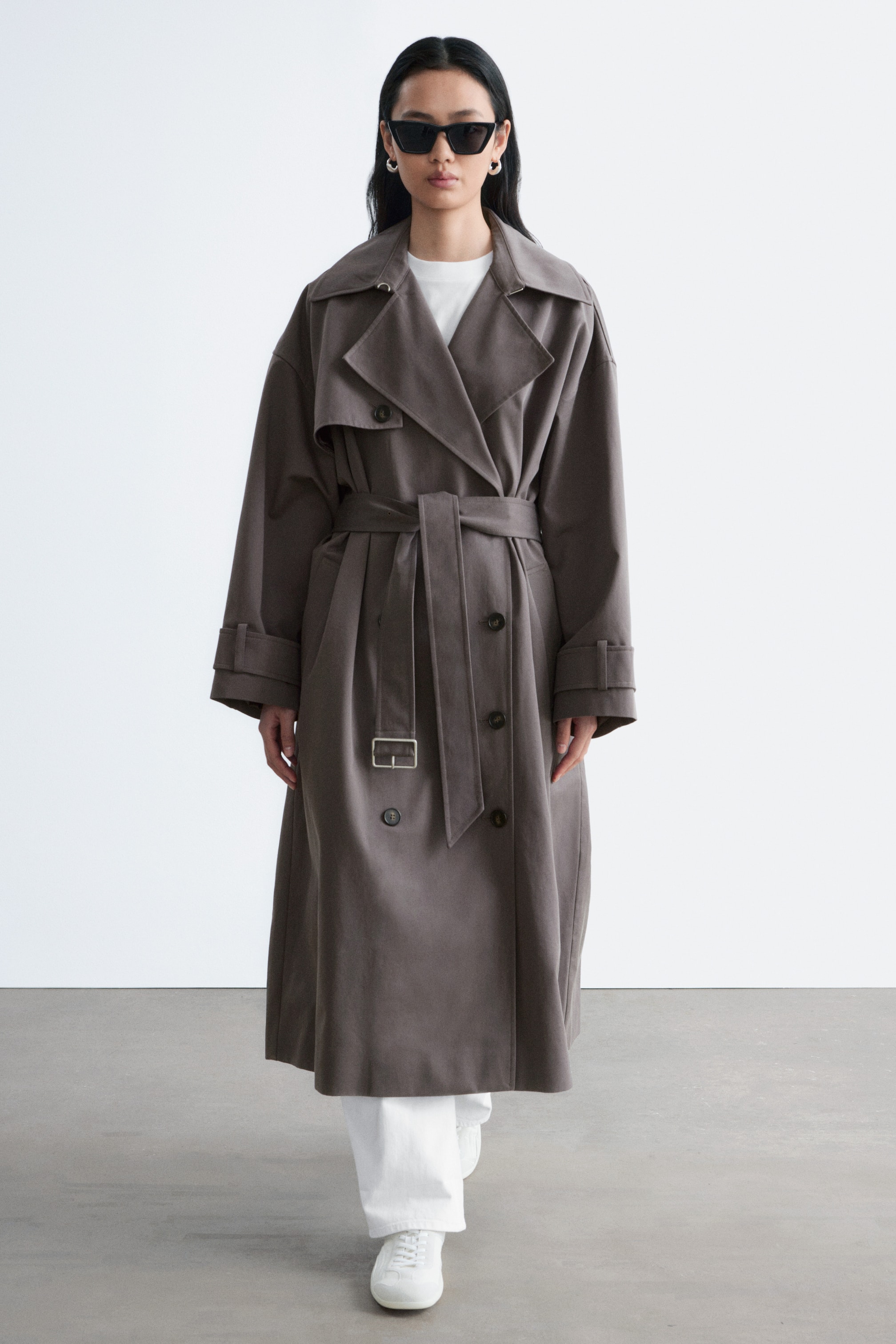 Nagyobb kép megtekintése: A modell egy sötétszürke, dupla soros ballonkabátot visel, amely széles hajtókás gallérral, hosszú ujjal és pántos részletekkel, valamint csattal ellátott szövetövvel van ellátva, amely karcsúsítja a derekat. A kabát egy fehér póló és fehér nadrág fölött van stílusosan viselve.