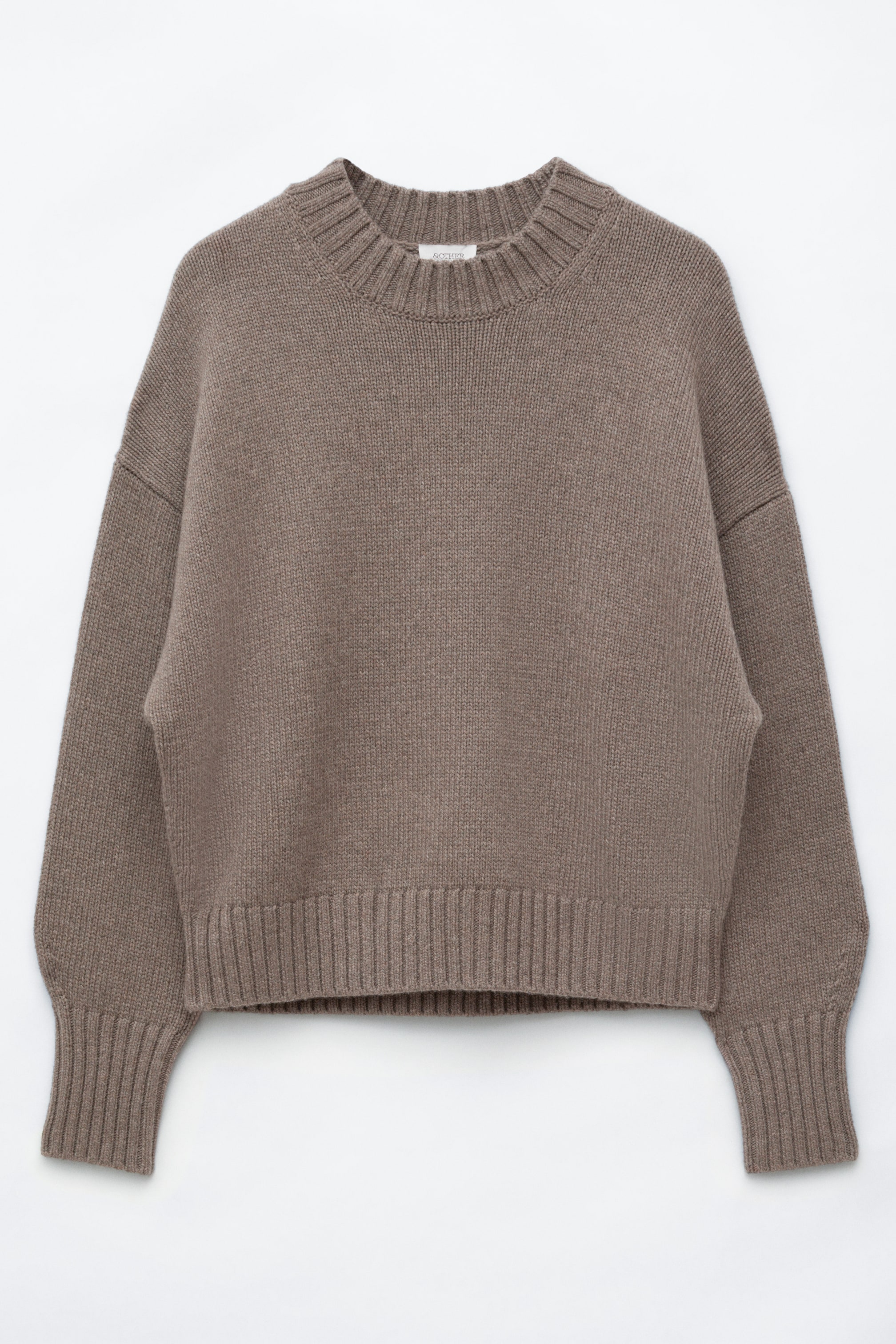 Visualizza immagine più grande: Un maglione in maglia color tortora è mostrato frontalmente, caratterizzato da una vestibilità rilassata e oversize con spalle scese e un ampio girocollo a costine spesse. Il modello presenta maniche lunghe rifinite con polsini a costine allungati e un ampio bordo a costine.